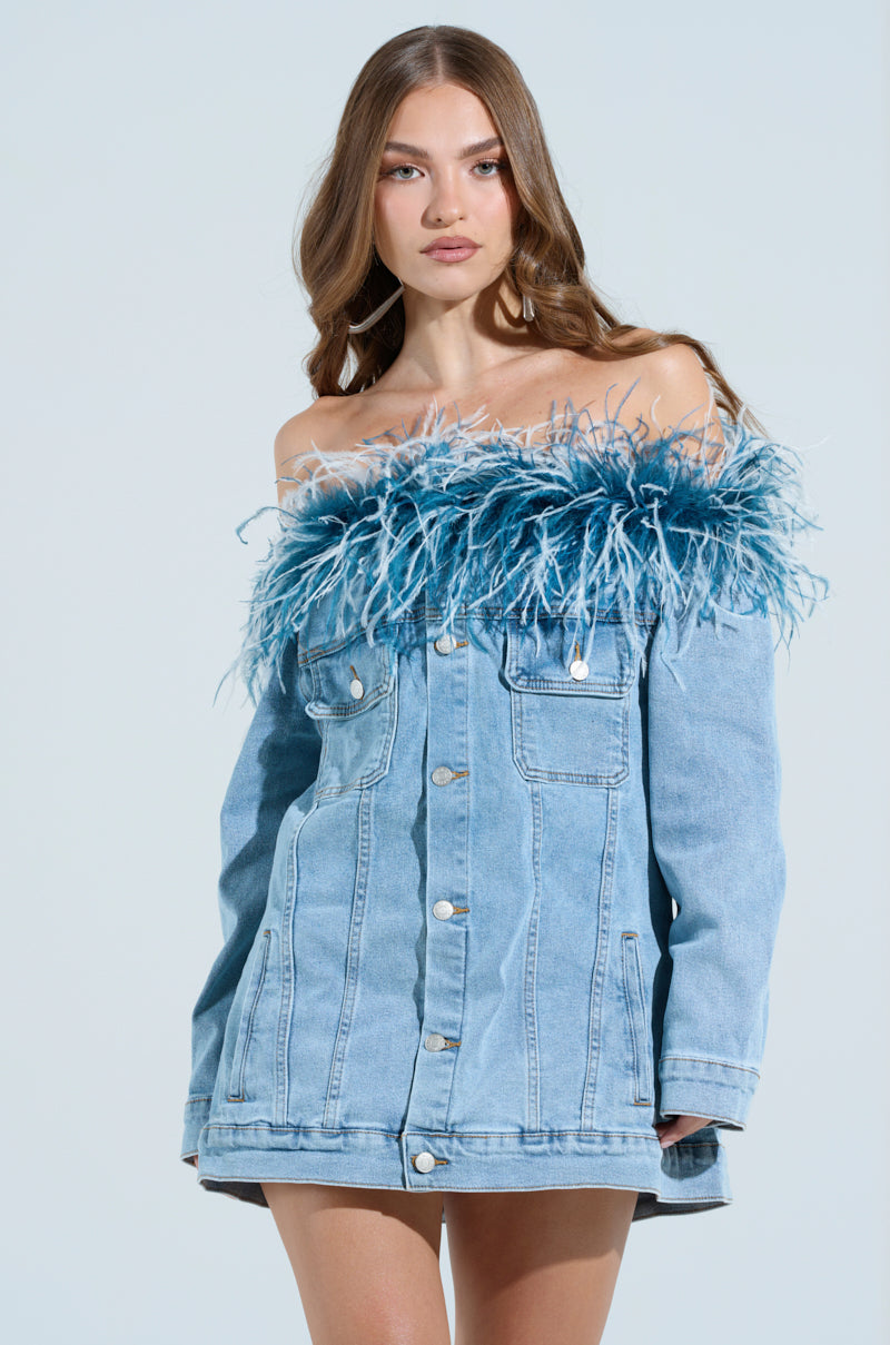 A woman poses in the CHIC AF DENIM MINI DRESS WITH FEATHERS. Her long brown hair cascades down against a plain background, showcasing her effortless style with an off-the-shoulder design and blue feathers along the neckline.