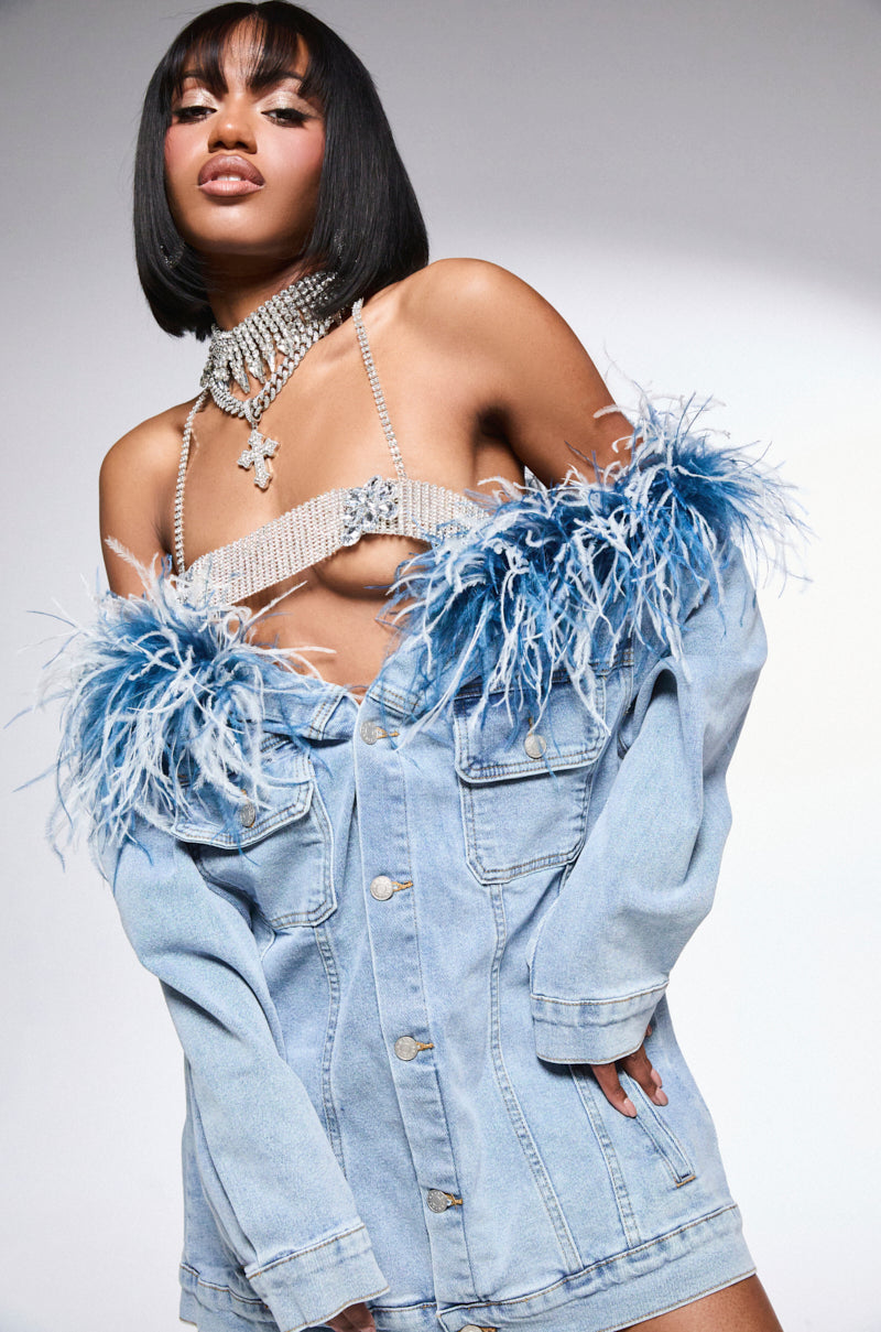 A woman confidently poses in a stylish outfit featuring the CHIC AF DENIM MINI DRESS WITH FEATHERS, complemented by a bejeweled top and a sleek bob hairstyle. The simple gray background highlights her fashion-forward look.