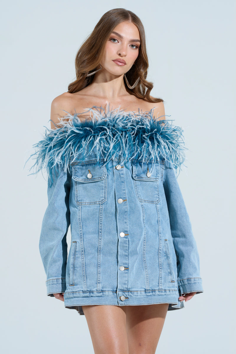 In a light blue setting, a woman models the CHIC AF DENIM MINI DRESS WITH FEATHERS featuring blue and white feather details. Her long brown hair flows elegantly, and she completes the chic look with hoop earrings.