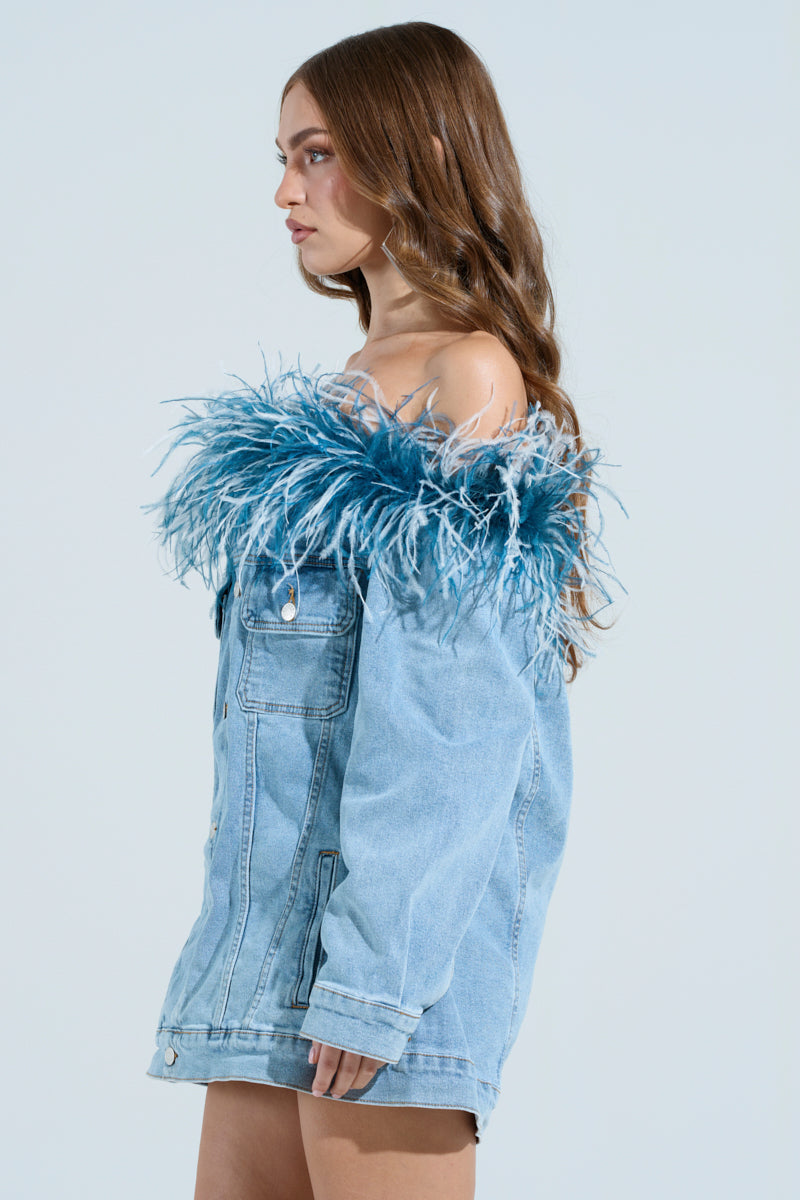A woman with long brown hair stands in profile, wearing the CHIC AF DENIM MINI DRESS WITH FEATHERS. The dress is an off-the-shoulder style, featuring blue and white feathers around the top. The background is plain light blue.