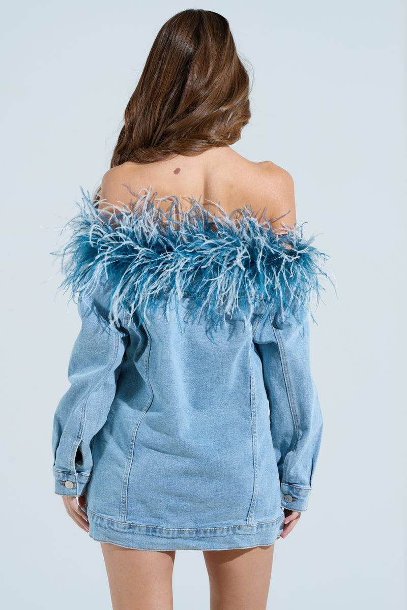 A woman with long brown hair wears the CHIC AF Denim Mini Dress with Feathers, featuring an off-the-shoulder design and feathery blue trim at the neckline. She faces away, showcasing the stylish back against a light blue background.