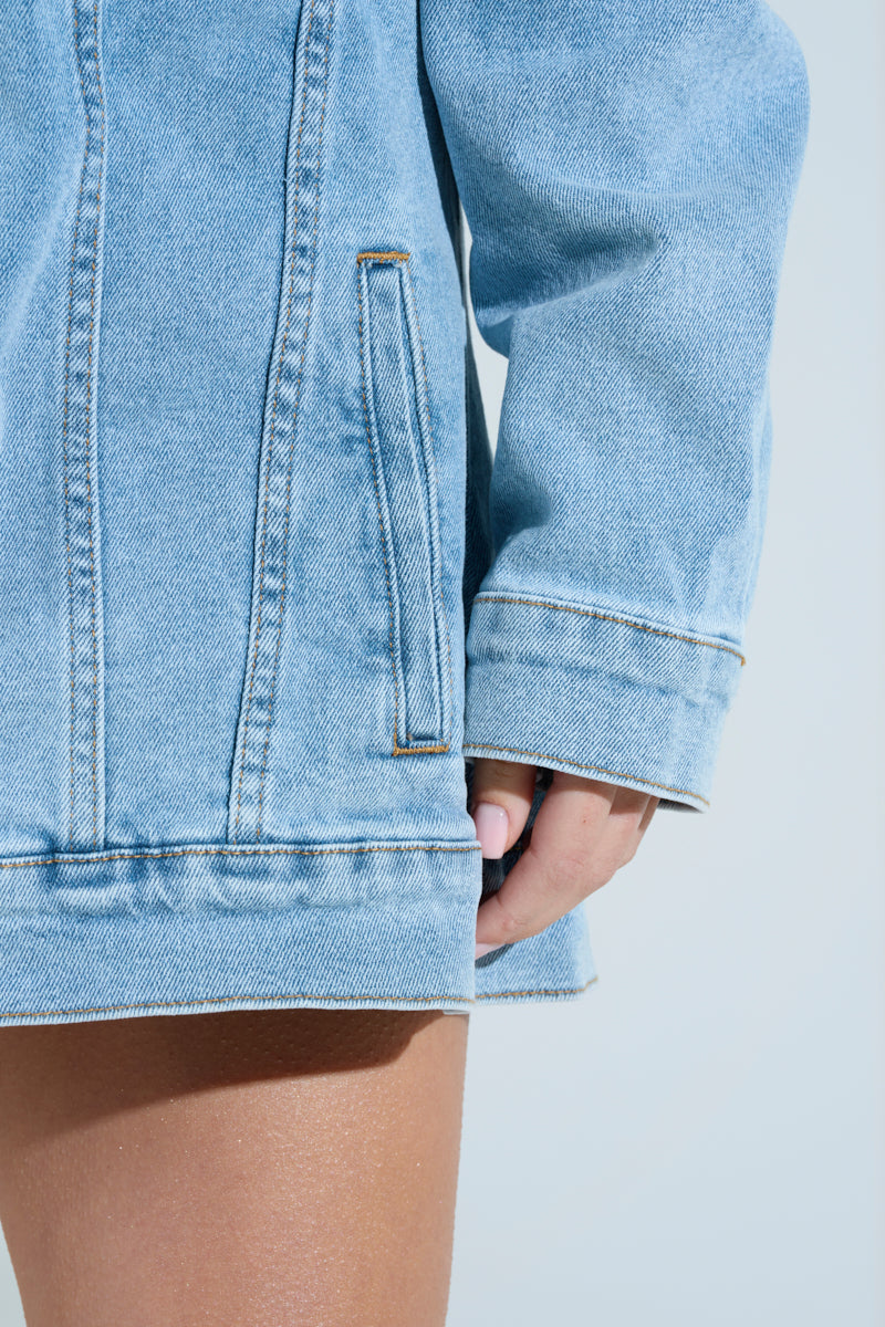 A person in the CHIC AF Denim Mini Dress with Feathers showcases this light blue dress, tucking a hand into a side pocket. Their bare leg peeks from beneath, emphasizing the denim's texture and stitching.