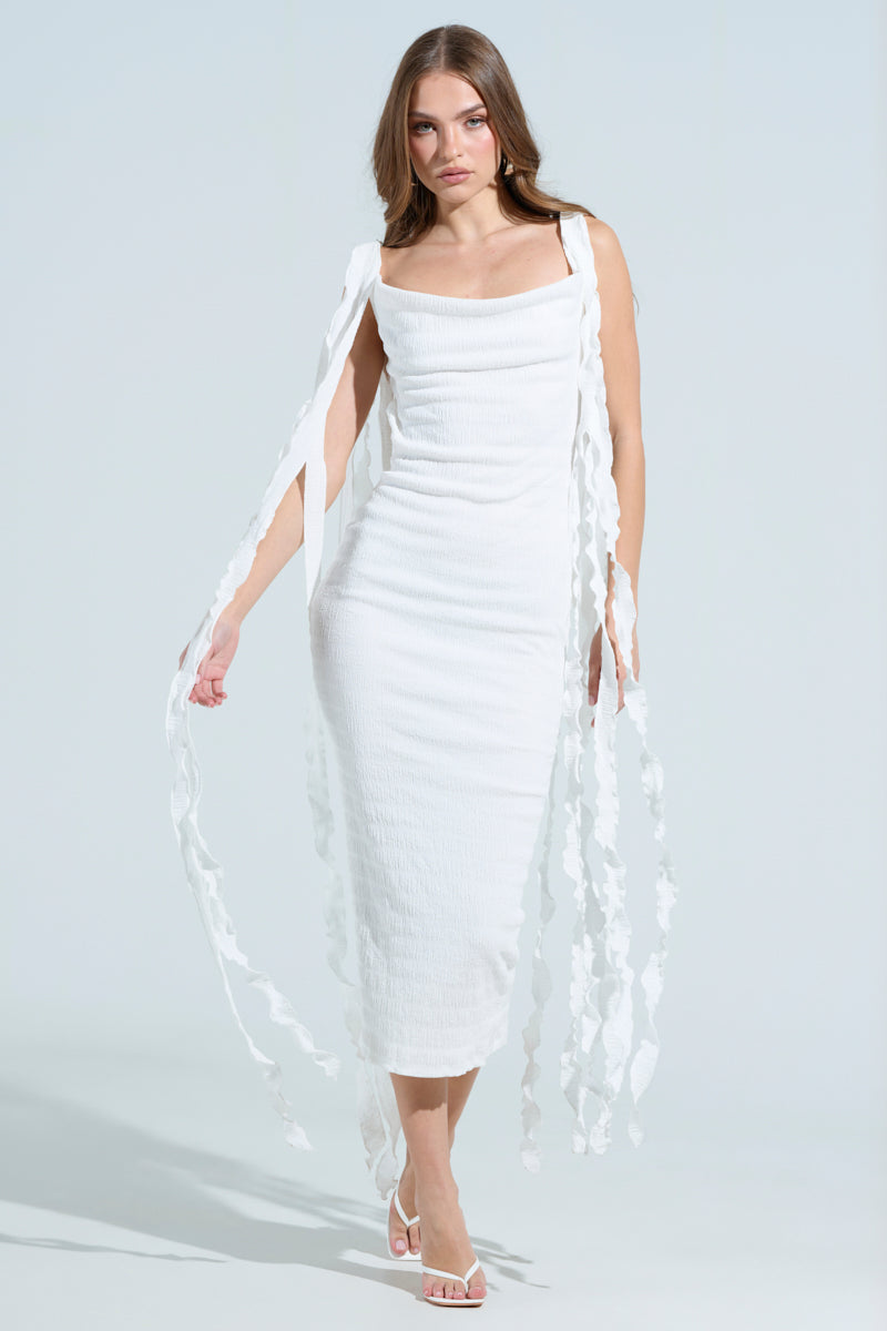 A woman wears the "GET LIKE ME BACKLESS MIDI DRESS IN IVORY," a sleeveless, backless midi dress with ribbon details on a light backdrop. The curve-hugging silhouette reaches her ankles, paired with white sandals, and her long hair flows gracefully.