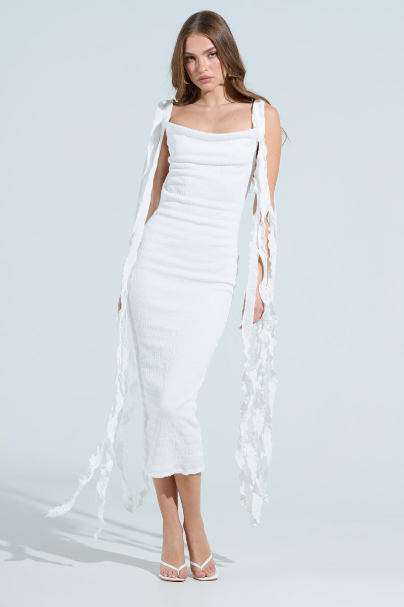 A woman poses against a plain background with long brown hair, wearing the curve-hugging GET LIKE ME BACKLESS MIDI DRESS IN IVORY with shoulder details extending into long ribbons. She completes her look with open-toed heels, placing one hand on her hip and the other by her side.
