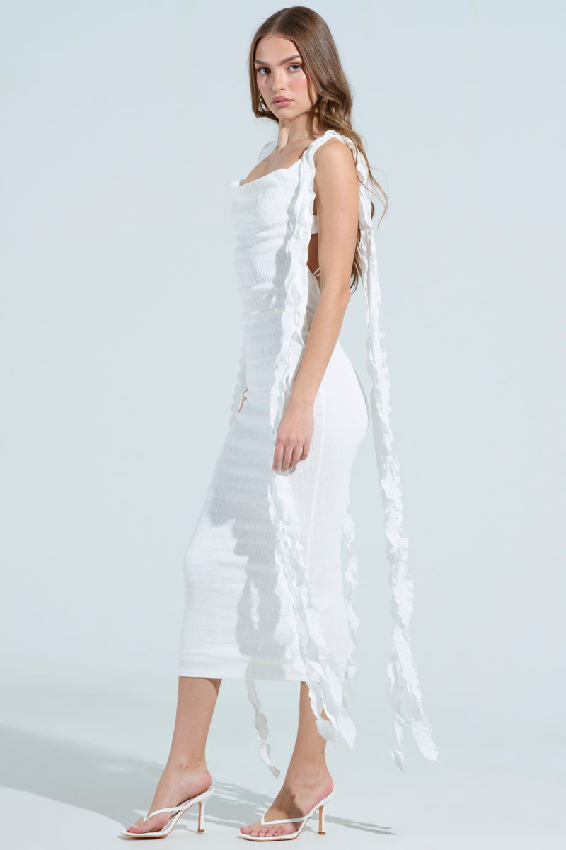 Against a light blue background, a woman in an ivory "GET LIKE ME BACKLESS MIDI DRESS" with long, flowing ribbons and white heels poses confidently. Her long hair cascades down her back, enhancing the curve-hugging silhouette of her dress.