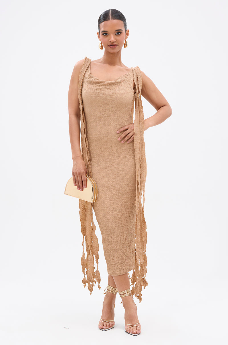 A woman poses against a white background wearing the GET LIKE ME BACKLESS MIDI DRESS IN MOCHA MOUSSE, a sleeveless, fitted beige midi with long fringe sides. She accessorizes with a gold clutch, strappy gold heels, and hoop earrings. 1