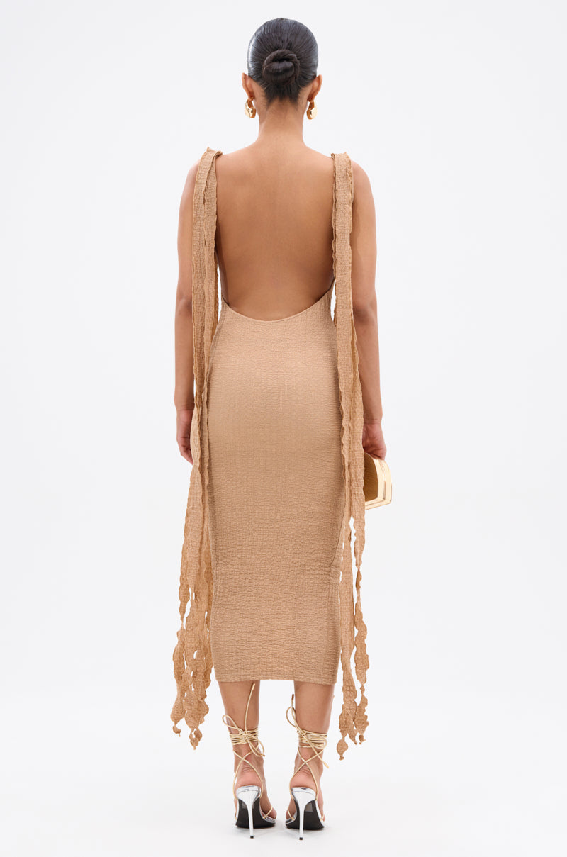 A woman faces away in the GET LIKE ME BACKLESS MIDI DRESS IN MOCHA MOUSSE—a curve-hugging, textured beige dress with a backless design and two long flowing panels. She styles her hair in a sleek bun, wears gold earrings, and carries a small handbag. 3