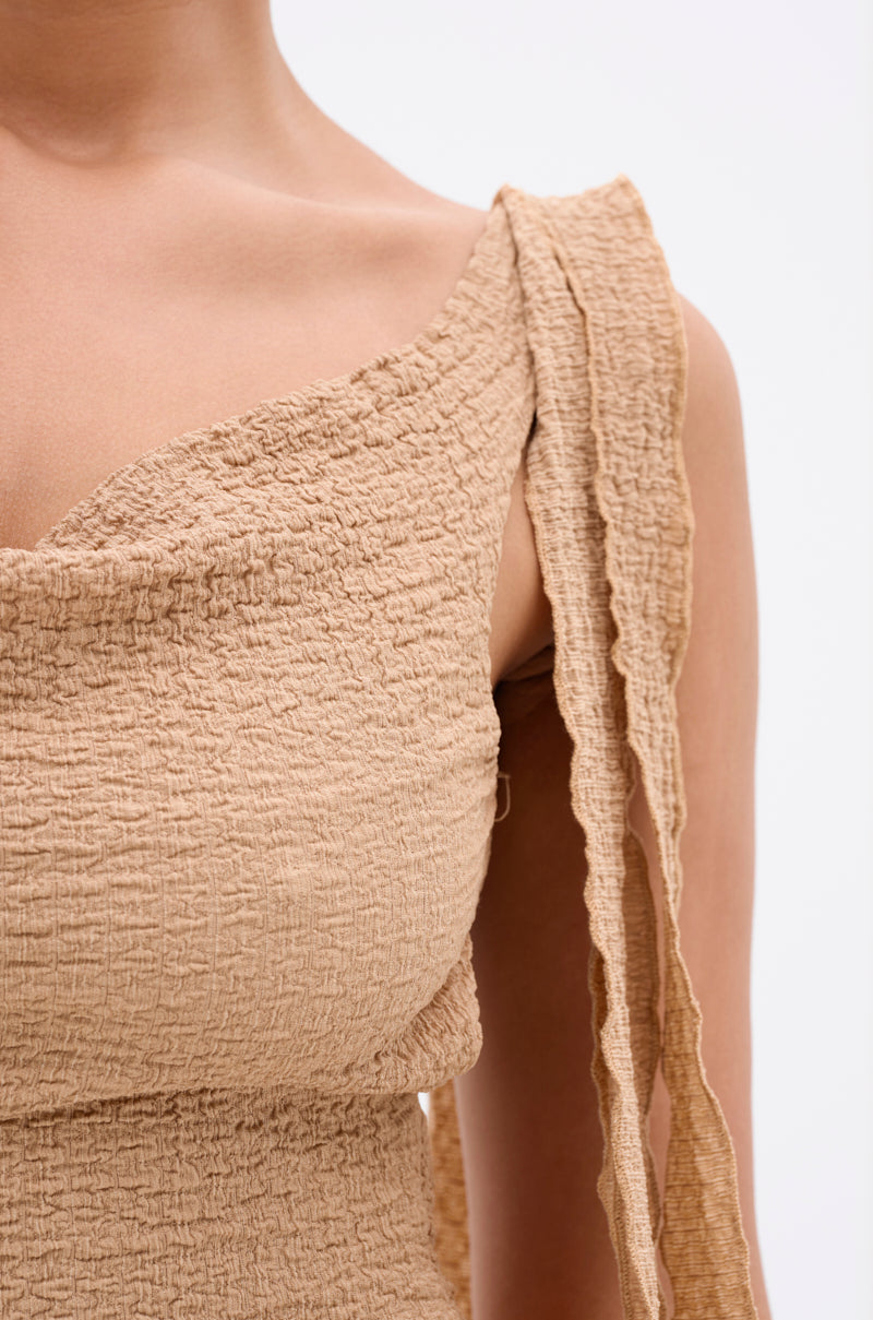 Close-up of a person wearing the GET LIKE ME BACKLESS MIDI DRESS IN MOCHA MOUSSE, featuring a textured, beige fabric with ruffled, one-shoulder strap and a crinkled, ruched finish for a curve-hugging silhouette. Plain light background. 4