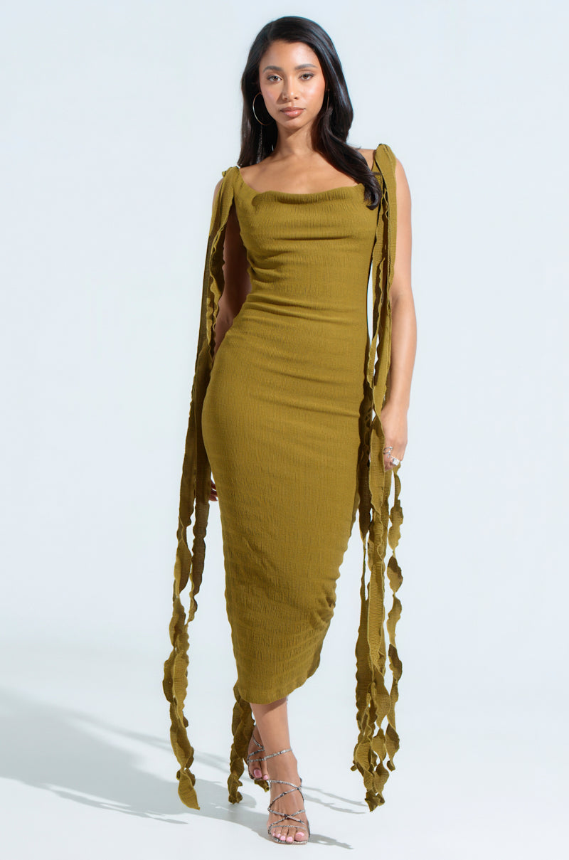 A woman poses confidently in a GET LIKE ME BACKLESS MIDI DRESS IN GREEN with shoulder ties and a body-hugging silhouette. The backless, olive green midi dress paired with strappy sandals stands out against the neutral background.