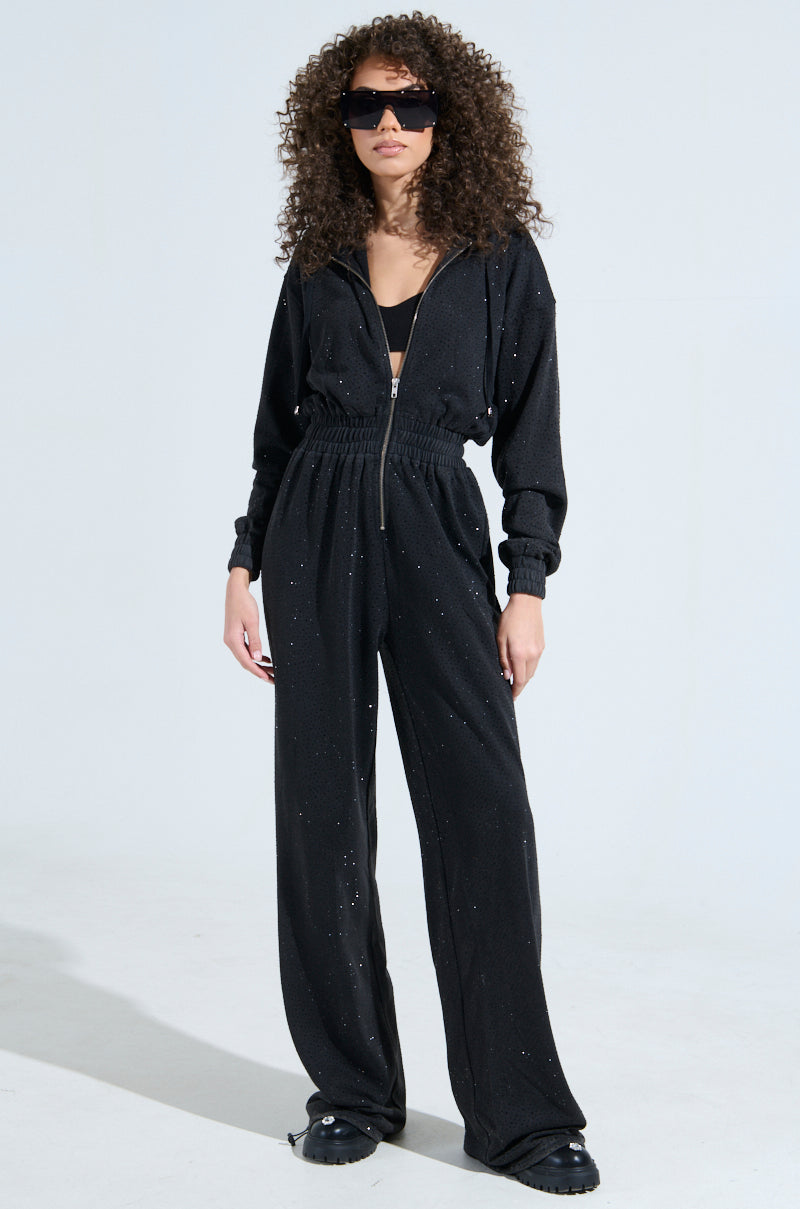 A person with curly hair confidently wears the DOING THE MOST French Terry jumpsuit featuring a metallic front zip closure, rhinestone embellishments, large sunglasses, and black shoes against a plain light gray background.