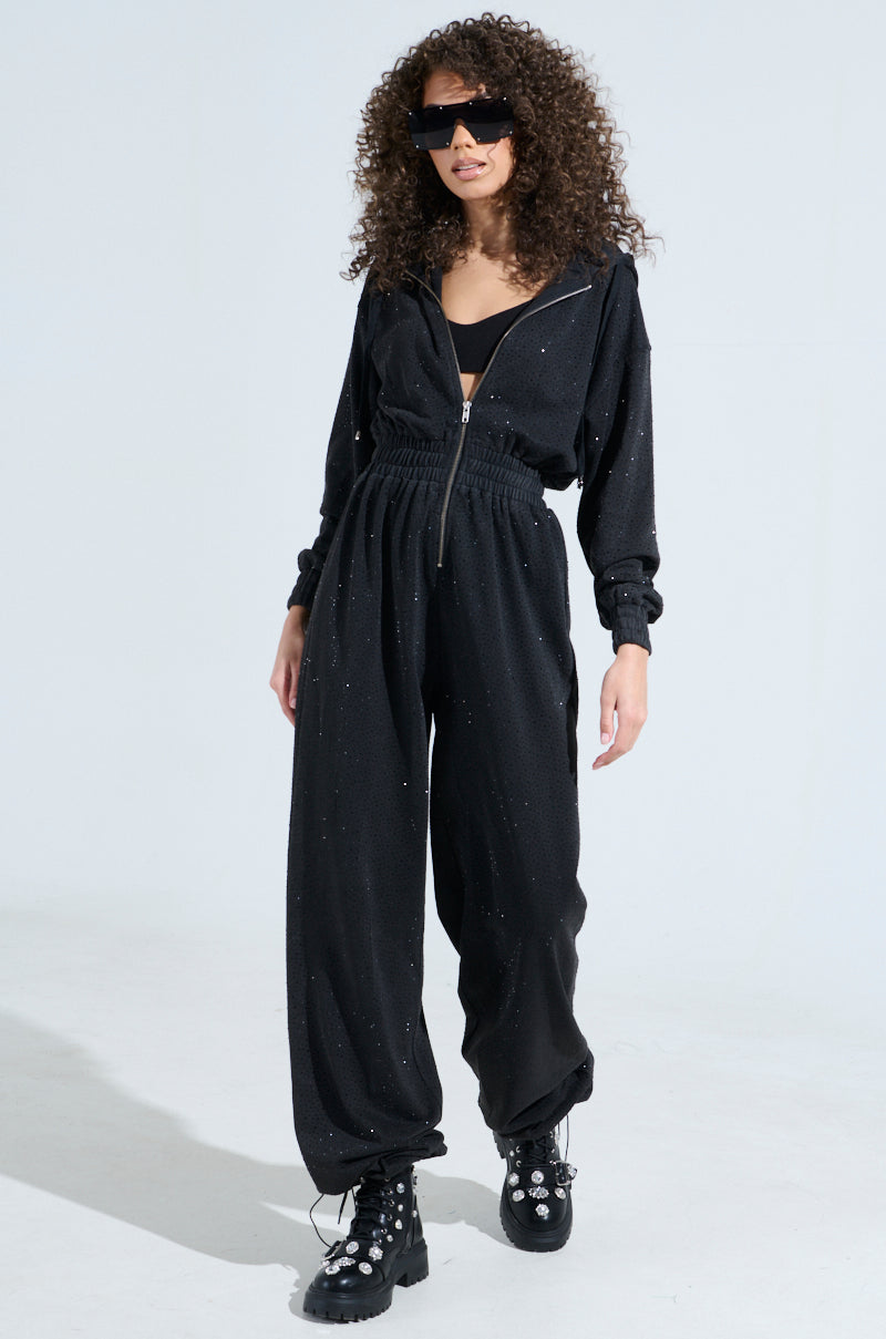 A person with curly hair wears the "Doing The Most French Terry Jumpsuit with Rhinestones," complemented by large sunglasses and black studded boots, as they confidently pose against a light background.