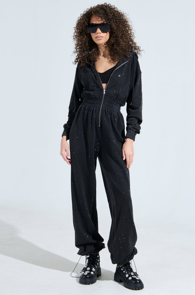 A person with curly hair exudes confidence in oversized sunglasses and the chic "Doing The Most French Terry Jumpsuit With Rhinestones," paired with black boots featuring decorative chains. The scene is set against a plain, light-colored background.