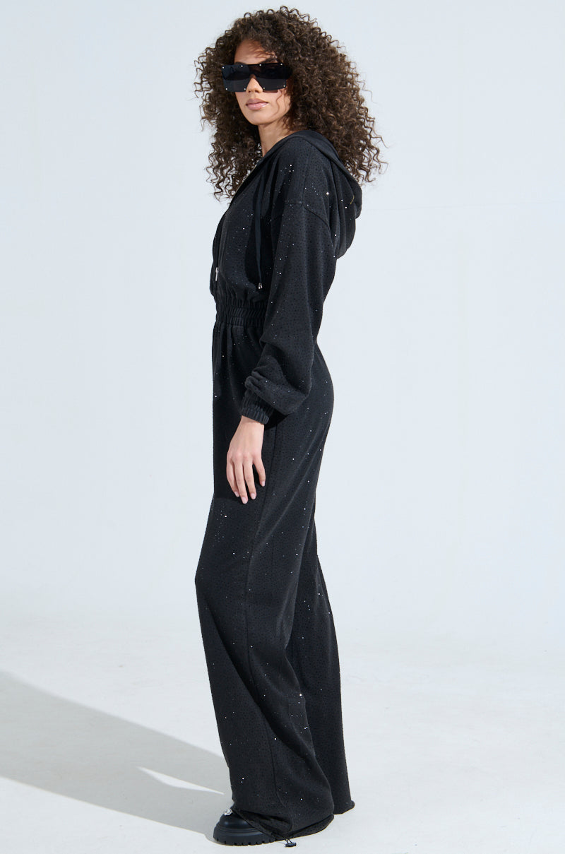 A person with curly hair and large sunglasses poses in the DOING THE MOST French Terry jumpsuit, featuring rhinestone embellishments, long sleeves, and a draped hood. They stand against a plain light background, gazing slightly to the side.