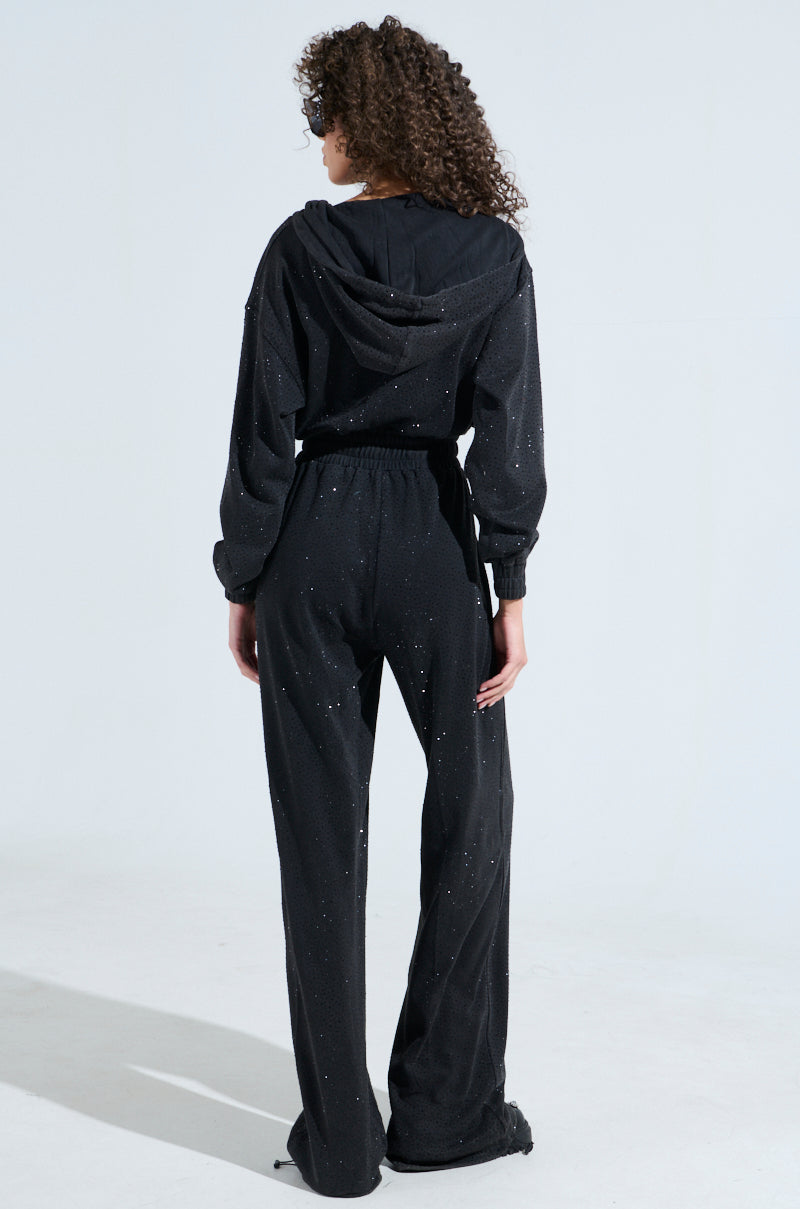 A person with curly hair is seen from the back, wearing the "Doing The Most French Terry Jumpsuit With Rhinestones" in black. They stand against a plain light background, hands by their sides, exuding effortless style and comfort.