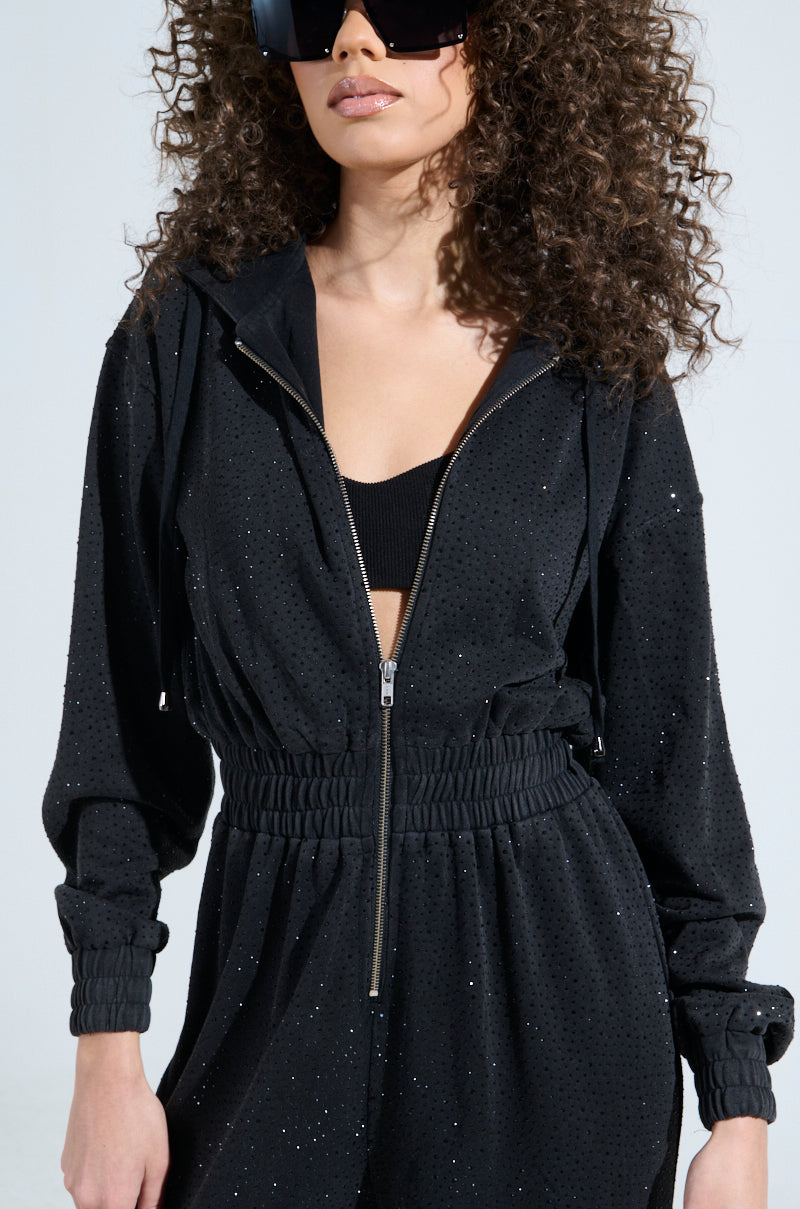 With curly hair and large sunglasses, a person rocks the DOING THE MOST FRENCH TERRY JUMPSUIT WITH RHINESTONES over a black top, set against a plain light background, highlighting their stylish presence.