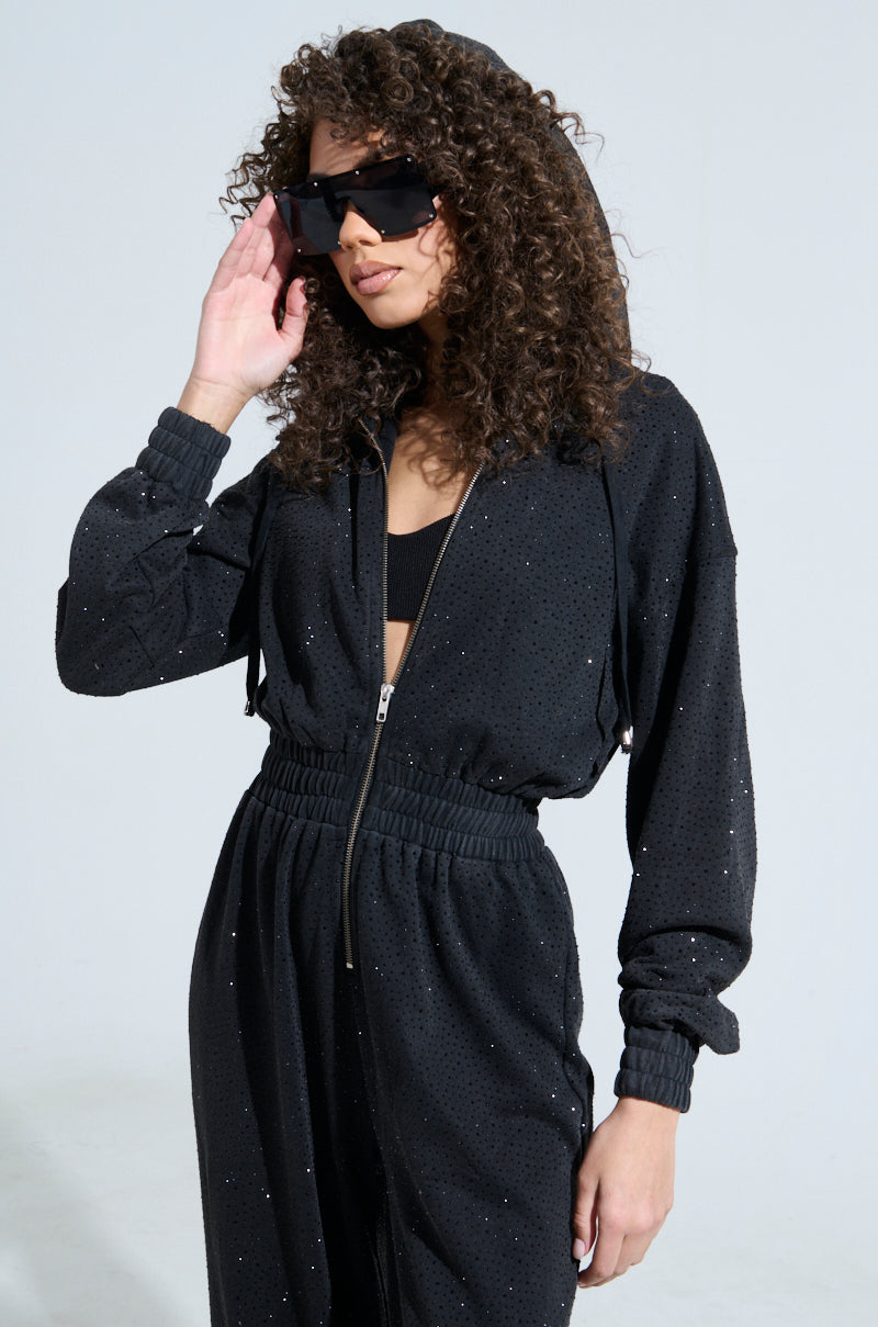 A person with curly hair and oversized sunglasses dazzles in the DOING THE MOST FRENCH TERRY JUMPSUIT WITH RHINESTONES, featuring a sparkly black design, rhinestone embellishments, and a metallic front zip. They're posed against a plain background, one hand casually touching their sunglasses.