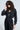 A person with curly hair and oversized sunglasses dazzles in the DOING THE MOST FRENCH TERRY JUMPSUIT WITH RHINESTONES, featuring a sparkly black design, rhinestone embellishments, and a metallic front zip. They're posed against a plain background, one hand casually touching their sunglasses.