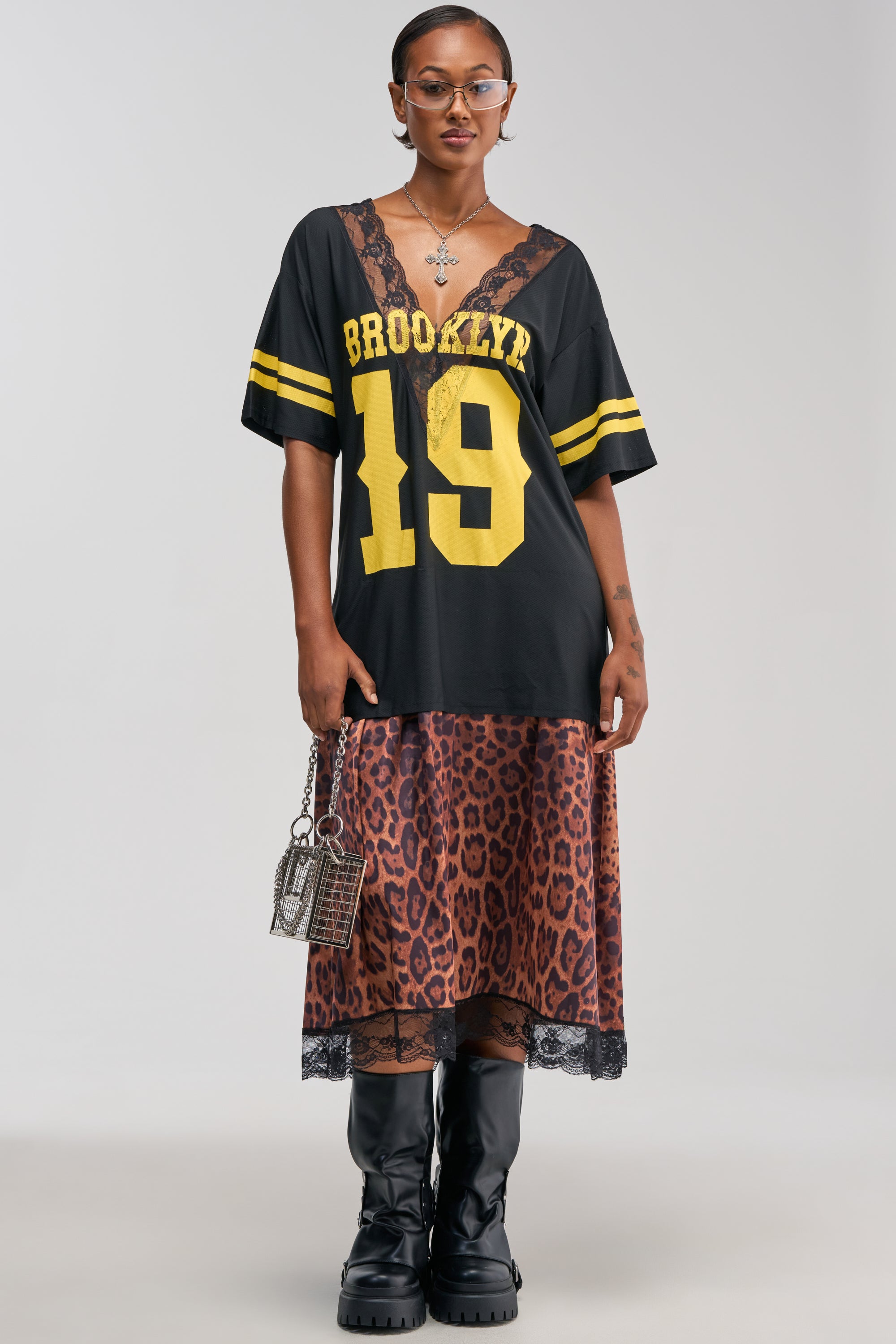 Sporting the CITY GIRL LAYERED JERSEY MAXI DRESS, this look features a chic black “Brooklyn 19” sports shirt over a cheetah print maxi dress, styled with black boots, clear glasses, hoop earrings, and a petite handbag—perfectly capturing effortless streetwear vibes against a simple backdrop.