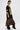 A woman stands sideways in the CITY GIRL LAYERED JERSEY MIDI DRESS, featuring a loose black jersey with “BROOKLYN 19” in yellow over a leopard print skirt. She accessorizes with hoop earrings and black heeled boots against a white background.