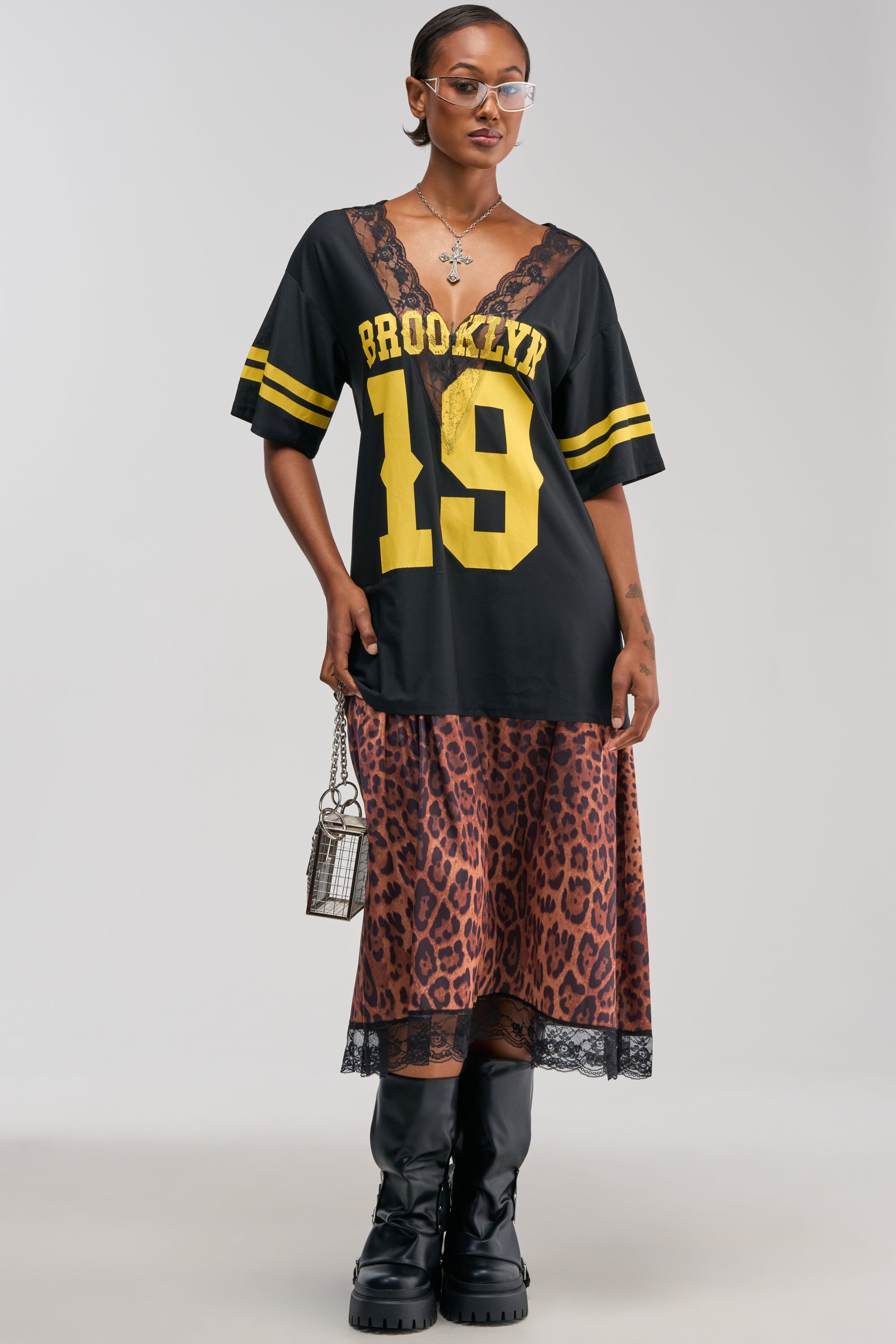 A woman wears the CITY GIRL LAYERED JERSEY MAXI DRESS in black, styled over a leopard print slip dress, paired with black knee-high boots, clear glasses, earrings, a necklace, and holds a small silver handbag.