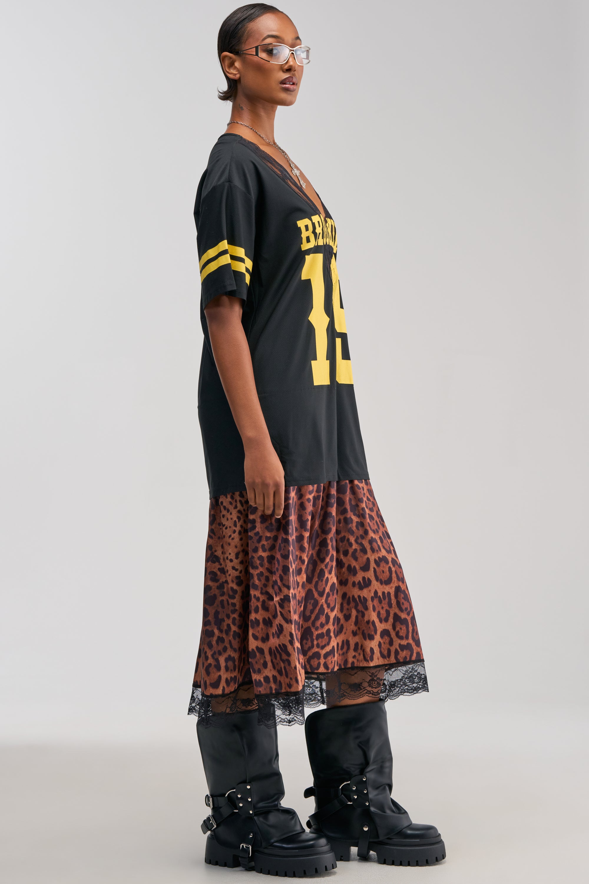 A person stands sideways against a plain light background, styled in the CITY GIRL LAYERED JERSEY MAXI DRESS—a black jersey with yellow stripes and the number 19, effortlessly layered over a cheetah print dress—completed with streetwear-inspired glasses and black knee-high buckled boots.