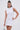 A woman with dark hair in a high bun wears the IDK FELT CUTE RIBBED KNIT MINI DRESS, a sleeveless white mock neck dress with a fitted silhouette. She holds a small silver handbag and wears large hoop earrings against a plain light background.