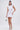 A woman poses against a plain backdrop in the IDK FELT CUTE RIBBED KNIT MINI DRESS, a sleeveless white mini dress. She pairs it with white heeled sandals, carries a silver handbag, and styles her long dark hair in a high ponytail.