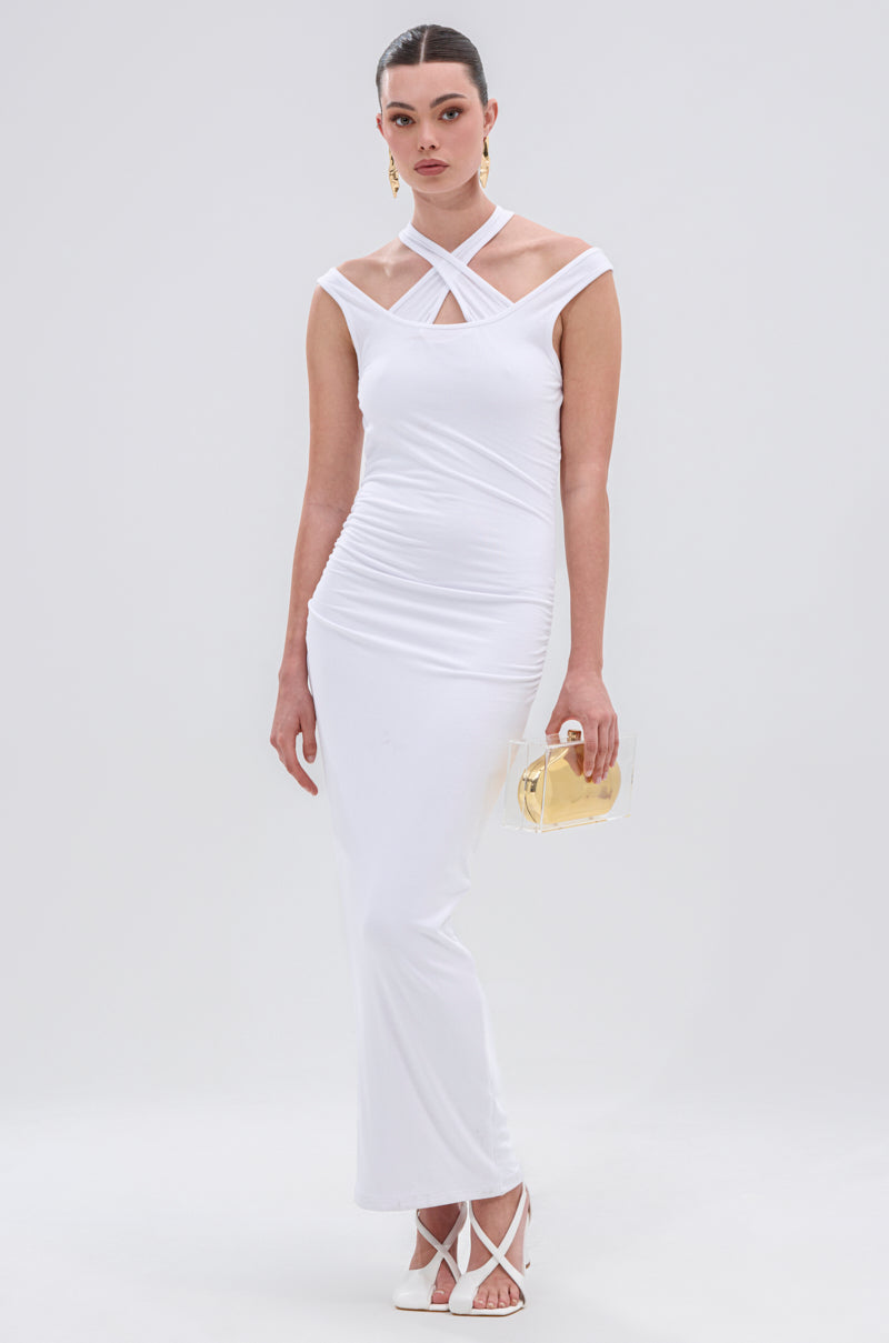 A woman wears the EXPRESS YOURSELF MAXI DRESS, a white fitted maxi with crisscross neckline straps and ruched details, paired with a clear handbag featuring gold accents and white strappy heels against a plain background.