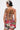 A person with short, dark hair and a platinum streak faces away, wearing the STREETS ARE CALLING PRINTED RUFFLE ROMPER with white lace-up strings and a graphic skirt. They display bold earrings and tattoos on their back and arm.