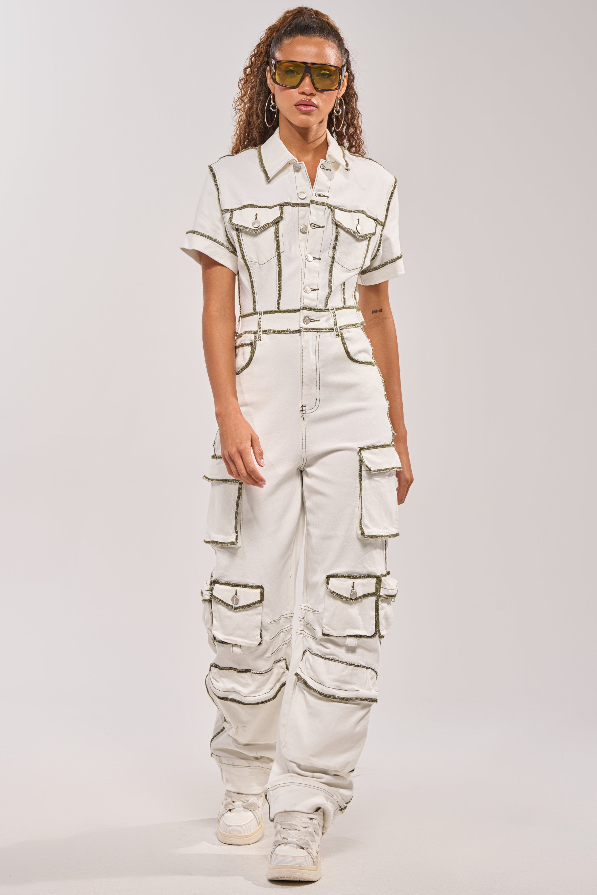 A woman wearing the BROOKLYN UTILITY DENIM JUMPSUIT with multiple cargo pockets and dark seams faces the camera. She pairs it with white sneakers and sunglasses, her curly hair framing her face against a plain, light background. 0