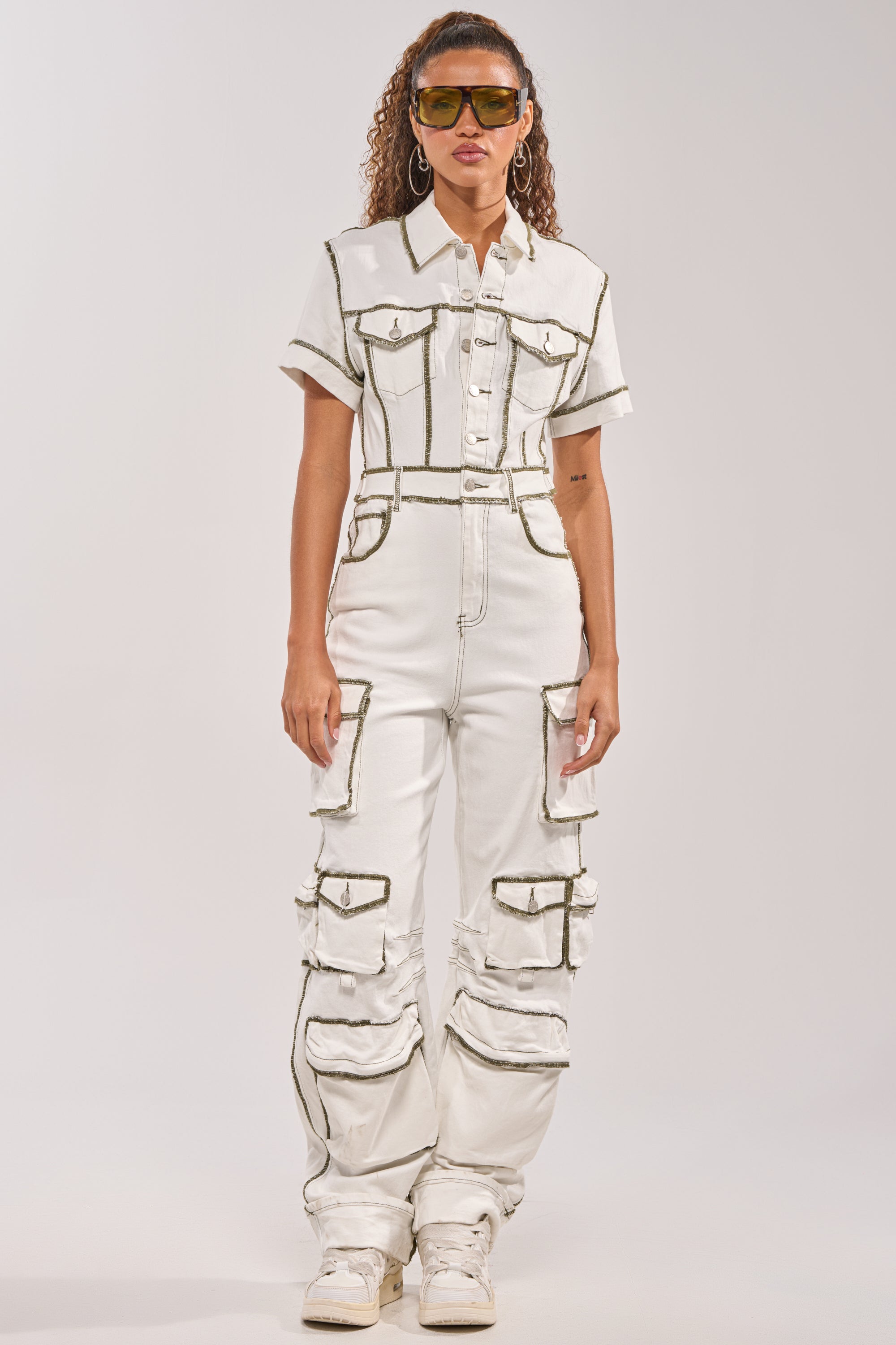 A woman stands facing forward in the BROOKLYN UTILITY DENIM JUMPSUIT with multiple pockets and dark seams, styled with white sneakers, large hoop earrings, and yellow-tinted sunglasses. 1