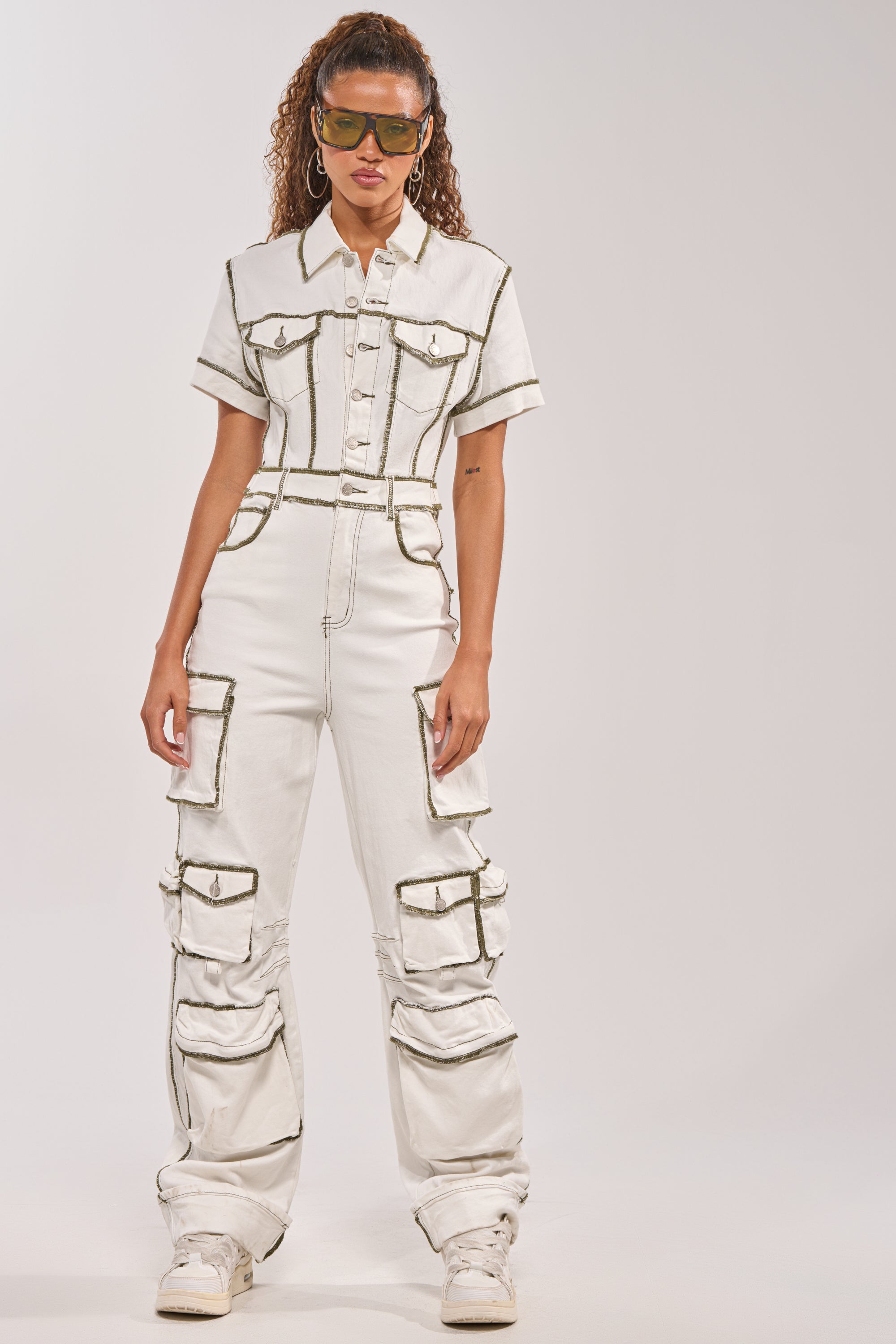 A person models the BROOKLYN UTILITY DENIM JUMPSUIT—white with dark seams, cargo pockets, short sleeves, and a belted waist—styled with white sneakers, large hoop earrings, and tinted sunglasses against a simple backdrop. 2