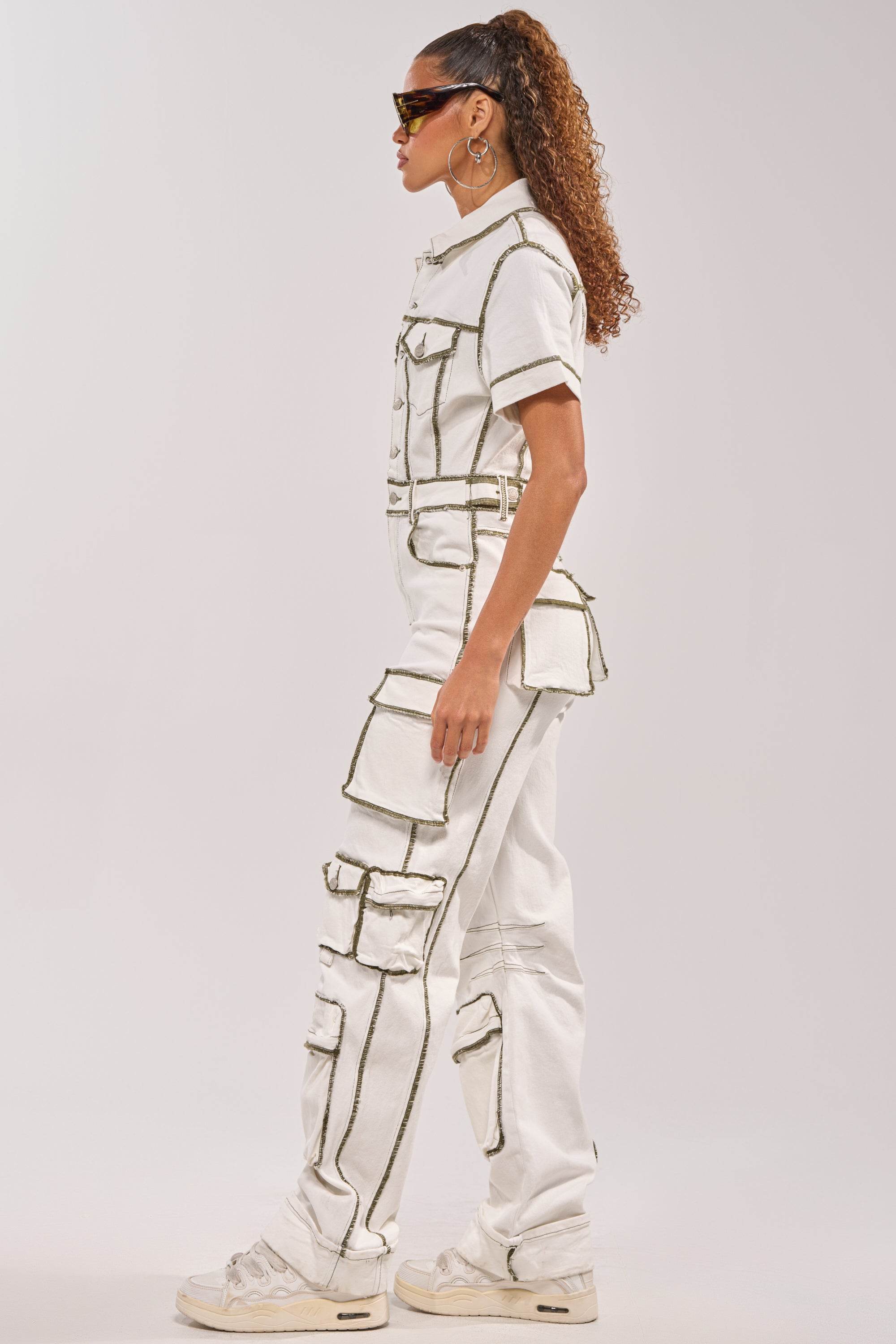 A woman in profile wears the BROOKLYN UTILITY DENIM JUMPSUIT, featuring multiple large pockets. She completes her look with chunky white sneakers, sunglasses, and a ponytail holding back her curly hair. 3