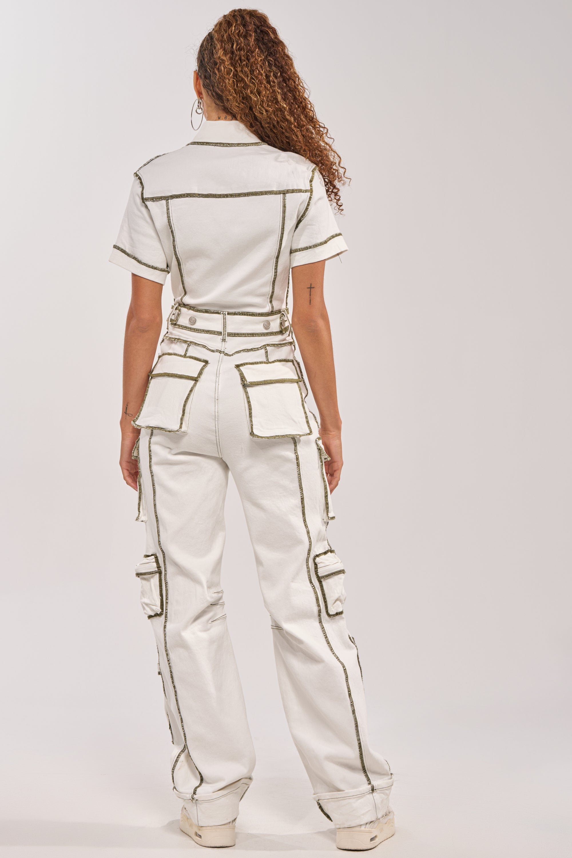 A person with curly hair faces away from the camera, wearing the BROOKLYN UTILITY DENIM JUMPSUIT—white with multiple pockets and bold, dark stitching—against a plain background. 4