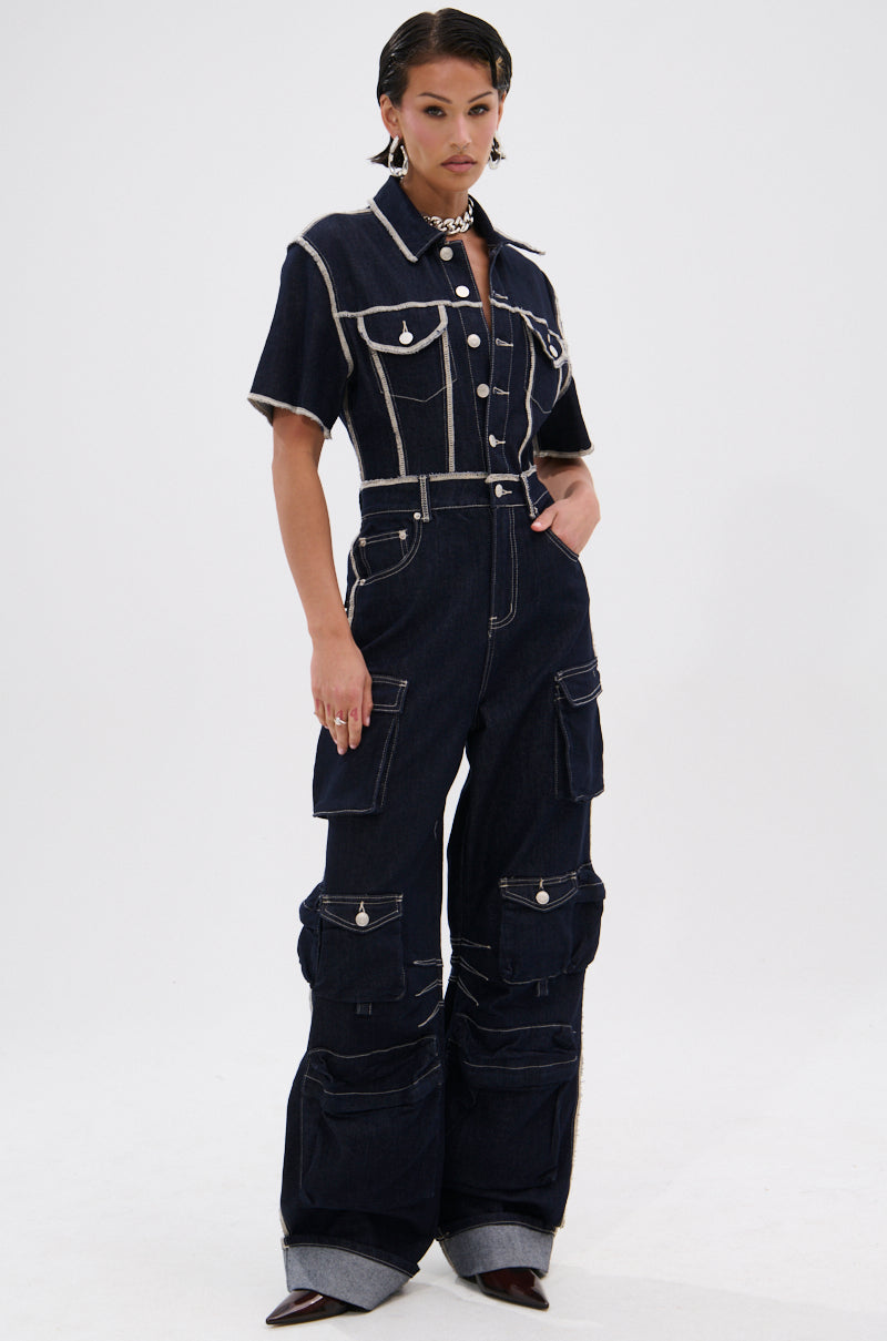 A person wears the ALEXANDRA DENIM CARGO JUMPSUIT with white contrast stitching, button-up front, cargo pockets, and wide cuffed legs. They style it with pointed shoes, short hair, bold makeup, and a chunky necklace.