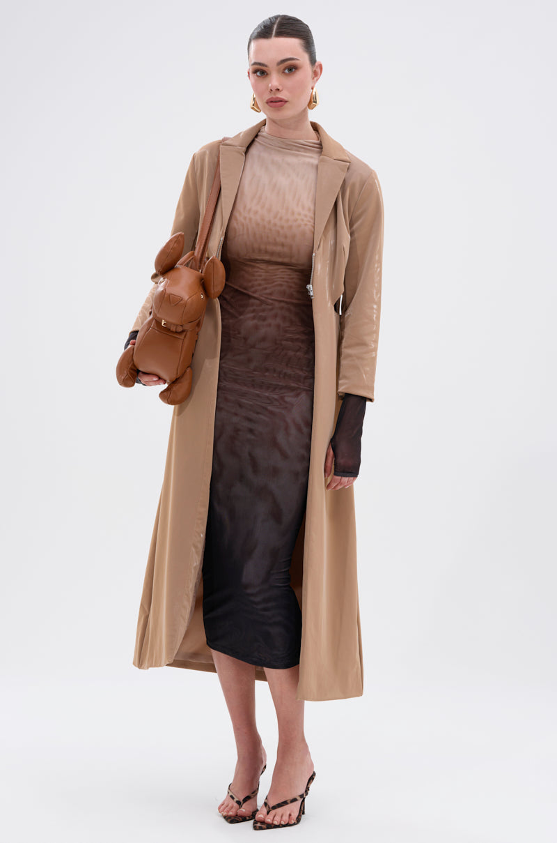 A woman stands against a white background wearing the FIFTY SHADES OMBRE MIDI DRESS under a beige trench coat, with strappy heeled sandals, gold earrings, and a tan dog-shaped handbag. Her hair is pulled back.