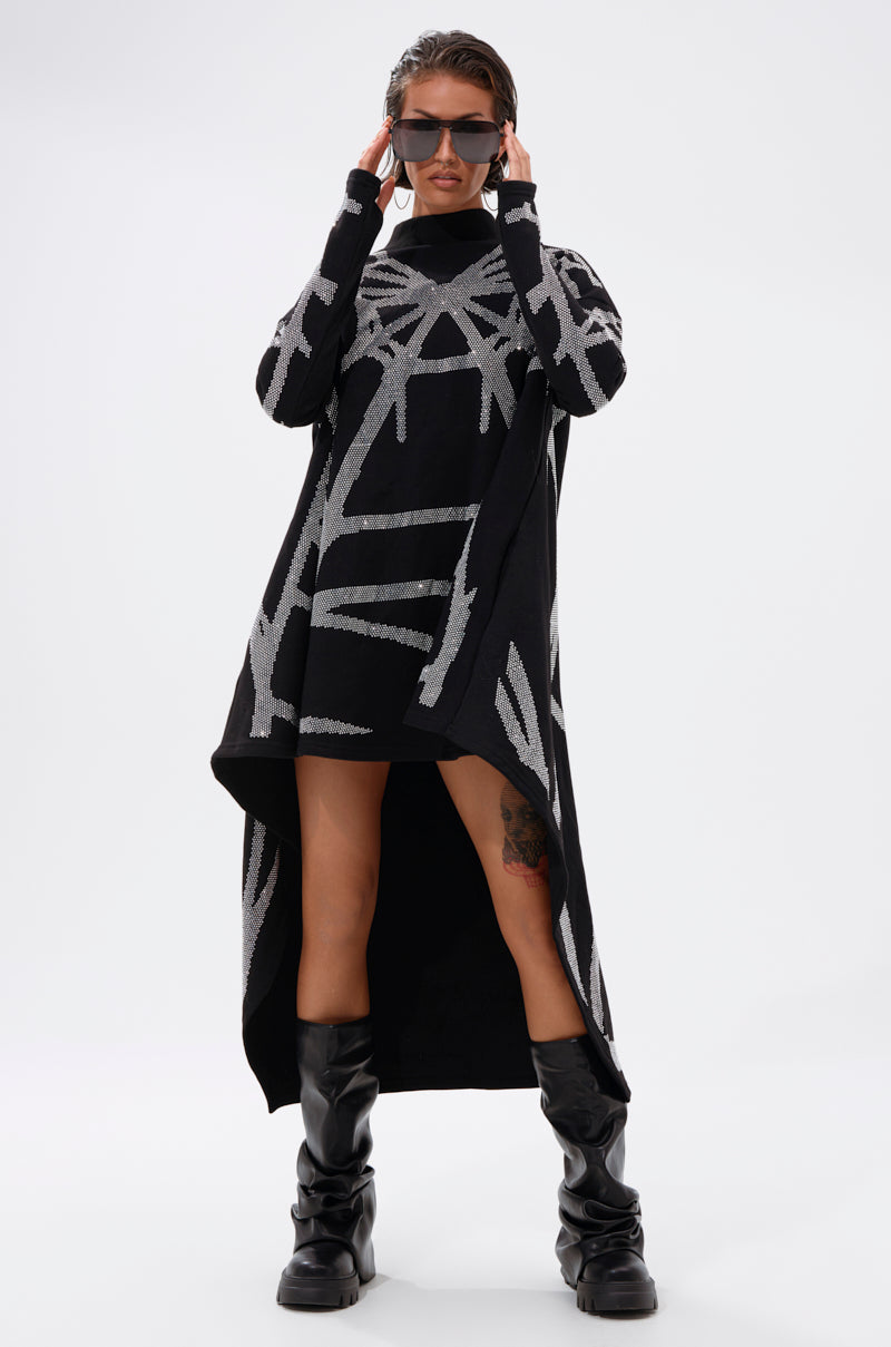A person in the LANIE PRINTED RHINESTONE SWEATER DRESS, featuring a black and gray asymmetrical hem with abstract lines, pairs it with black sunglasses and tall black boots, standing confidently against a white background.