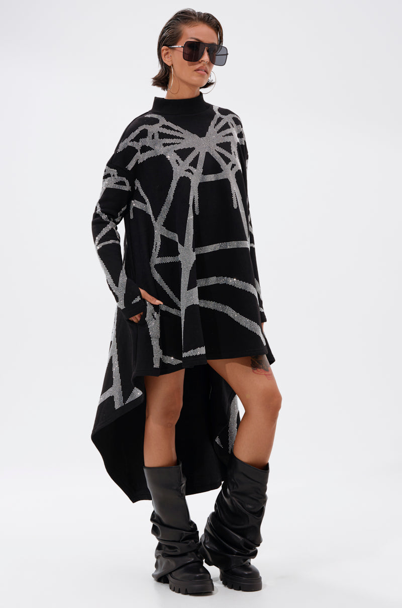 A person stands against a white background wearing oversized sunglasses, slouchy black knee-high boots, and the LANIE PRINTED RHINESTONE SWEATER DRESS with an abstract pattern and asymmetrical hem.