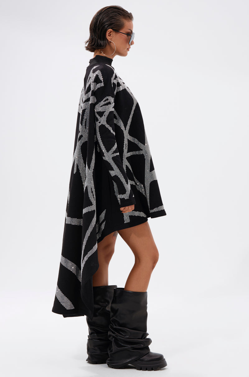 Wearing sunglasses, a loose black cape with silver patterns over the LANIE PRINTED RHINESTONE SWEATER DRESS, and oversized black slouchy boots, a person stands sideways against a plain light background.