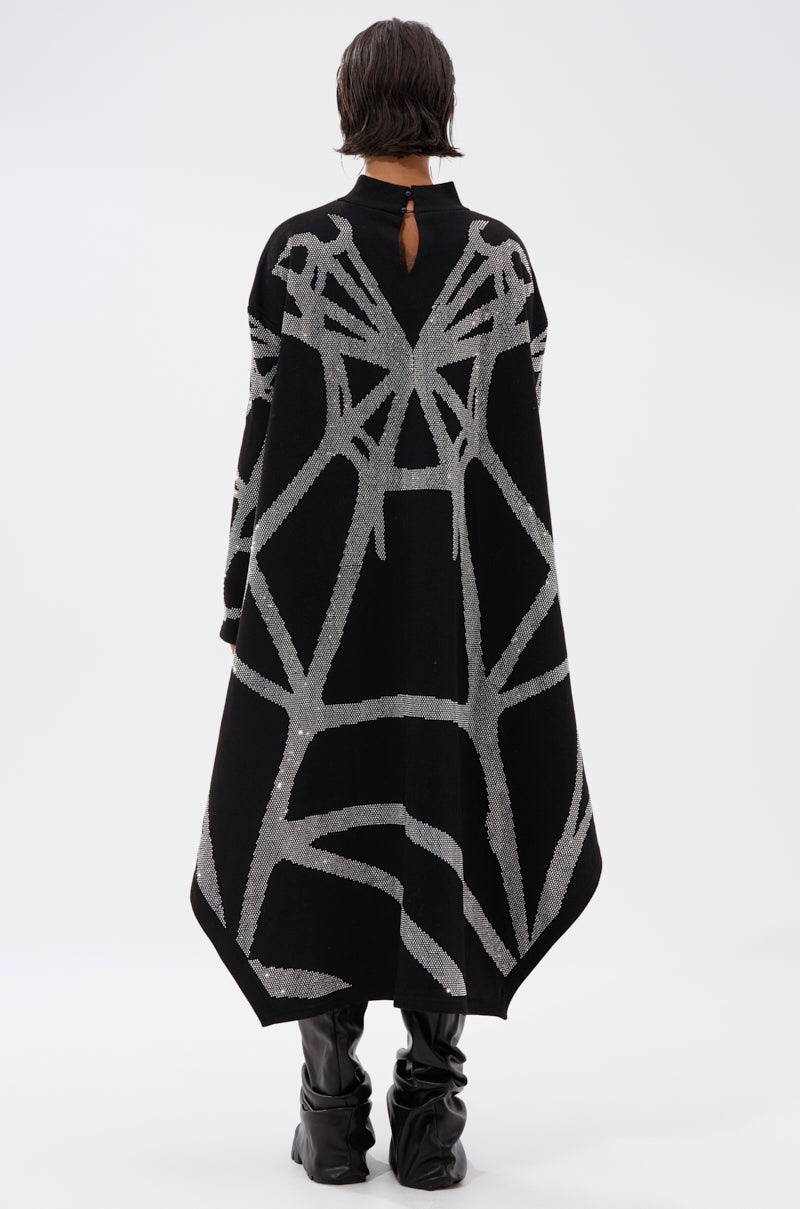 Seen from behind against a plain white background, a person with short dark hair wears black knee-high boots and the LANIE PRINTED RHINESTONE SWEATER DRESS, styled with a long black cape featuring bold silver geometric patterns.