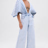 A person with short dark hair stands confidently against a white background, wearing the BORN TO STAND OUT DENIM JUMPSUIT featuring a plunging V-neckline and wide-leg silhouette.