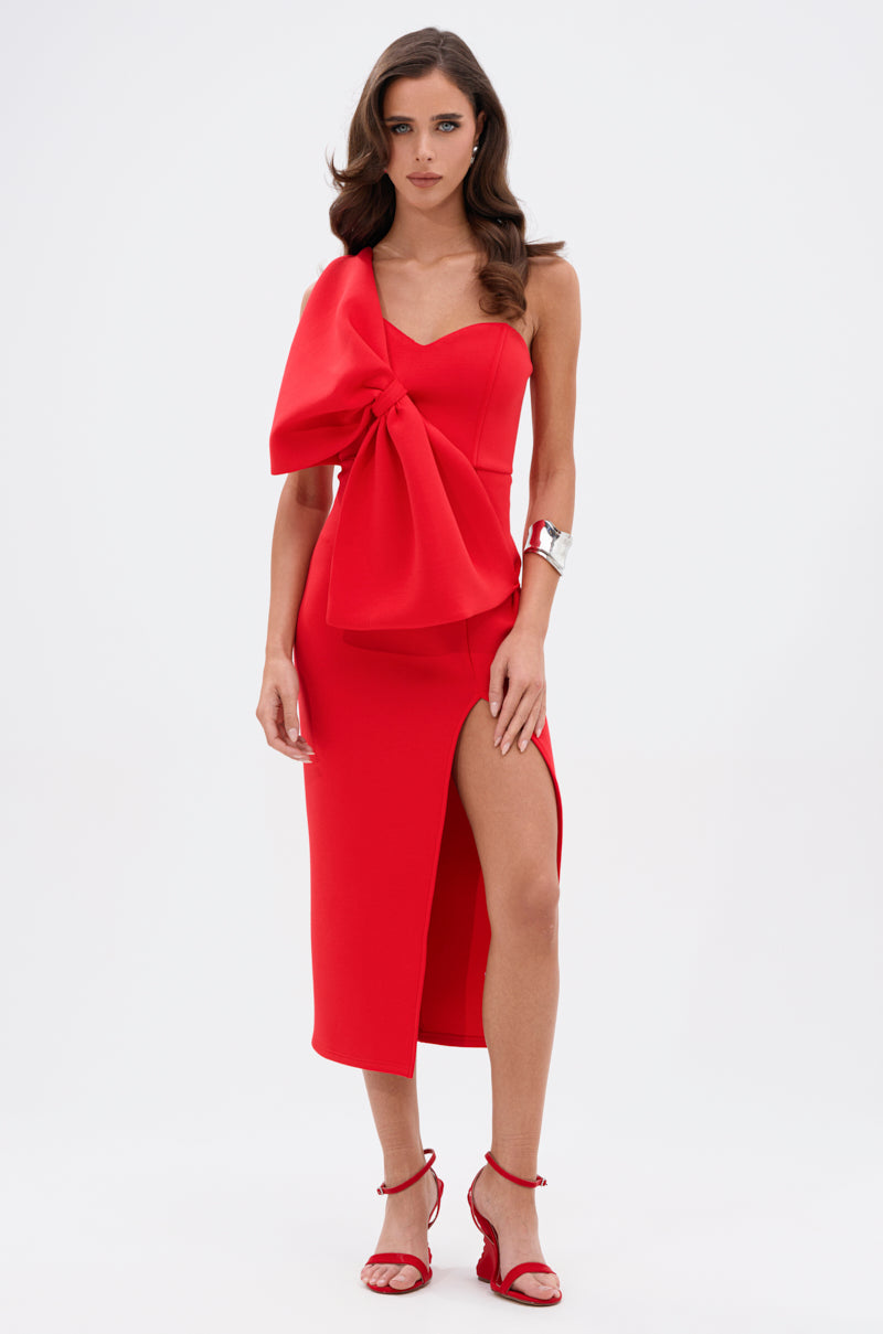 A woman stands confidently in the I WIN BOW FRONT MIDI DRESS, featuring a bold red hue, one-shoulder design, and thigh-high slit, paired with red heels and a metallic cuff bracelet against a plain white background.
