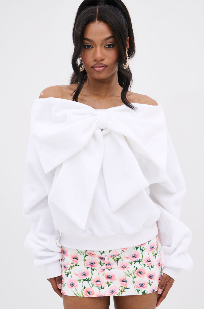 Woman wearing THE BEST BOW SWEATER, an off-the-shoulder white top with a bow applique, paired with a short pink floral skirt, standing against a plain light background.