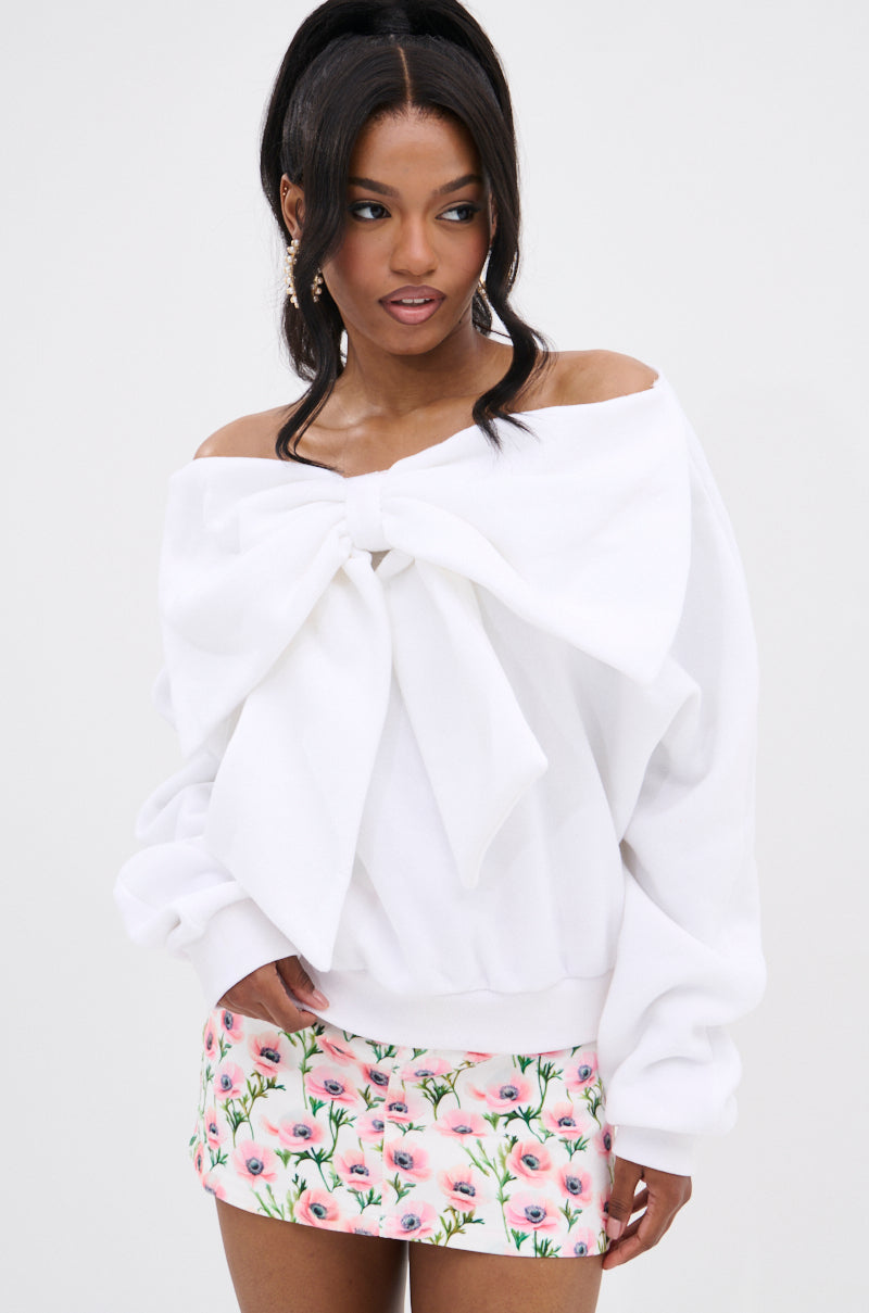 A woman wears THE BEST BOW SWEATER, a cozy off-the-shoulder top with a bow applique, paired with a short pink floral skirt, as she stands against a plain light background looking to the side.