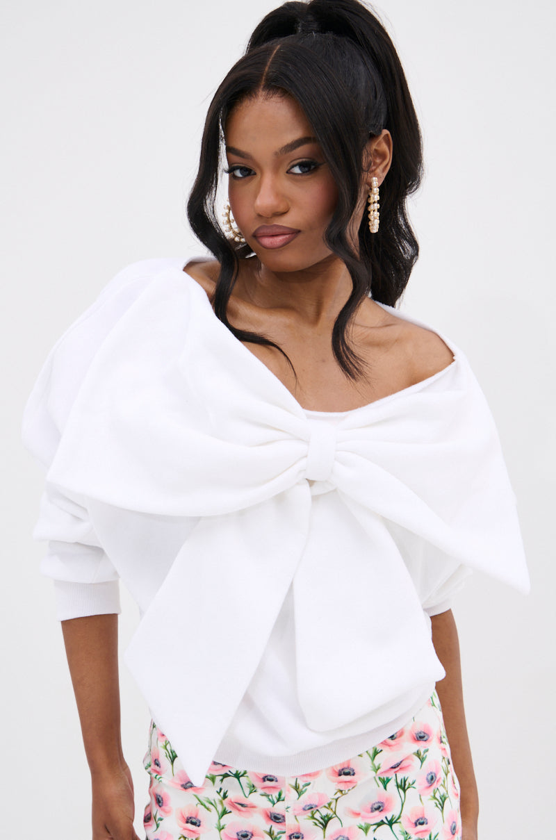 A woman with long dark hair in a high ponytail wears THE BEST BOW SWEATER, a white off-shoulder top featuring a large bow, paired with pink floral pants and statement earrings, posing against a plain light background.