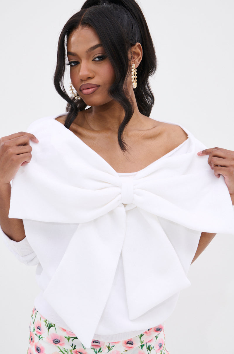 A woman with long dark hair wears THE BEST BOW SWEATER, a white oversized top with a bow applique, paired with pearl earrings and a white skirt featuring a pink floral pattern. She holds the sides of the bow and looks slightly to the side.