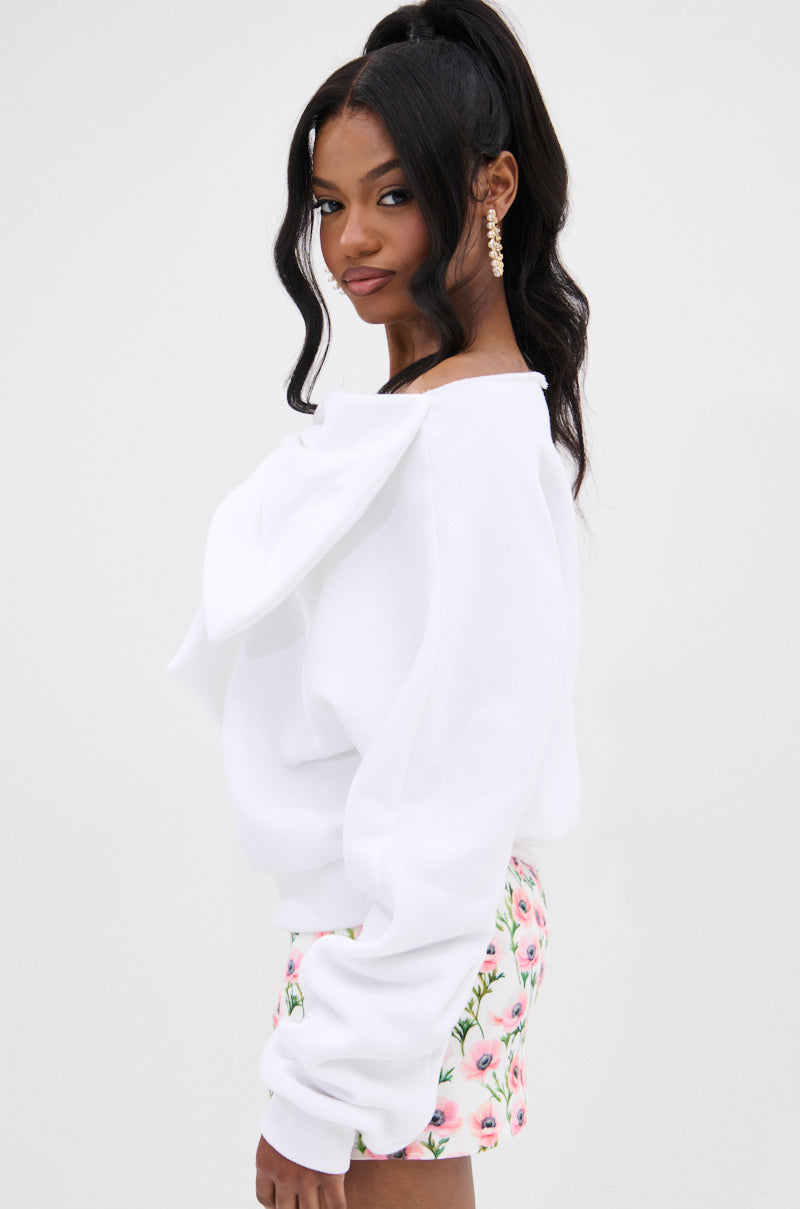 A woman with long black hair in a high ponytail models THE BEST BOW SWEATER, featuring a large bow appliqué, and poses sideways with a smile against a plain white background.