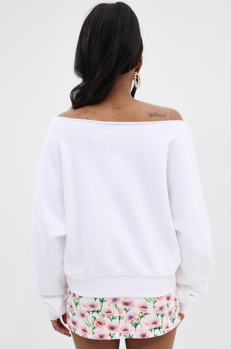 A woman with long dark hair and gold hoop earrings models THE BEST BOW SWEATER, a cozy off-the-shoulder knit styled with a pink floral mini skirt. She stands against a plain white background, showcasing an elegant oversized silhouette and a back tattoo.