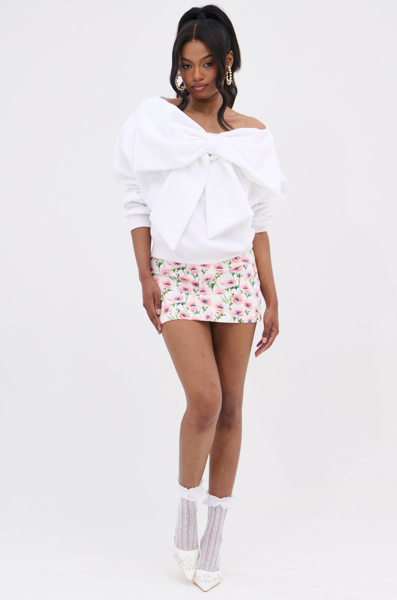 Woman wearing THE BEST BOW SWEATER, a white top with a large bow applique, paired with a pink floral mini skirt, white lace socks, pointed heels, and statement earrings against a plain white background.