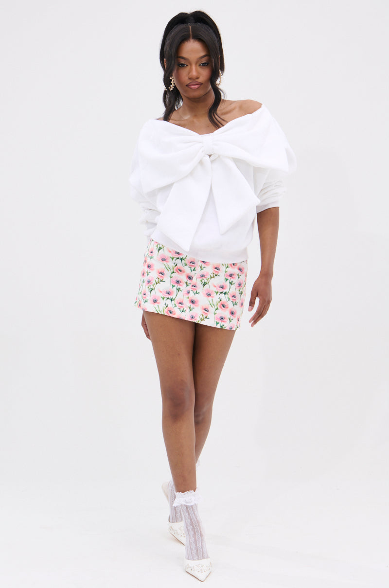 A woman with long dark hair wears THE BEST BOW SWEATER, an off-the-shoulder white top with a large bow applique, styled with a pink floral mini skirt, white lace socks, white heels, and statement earrings against a plain white background.